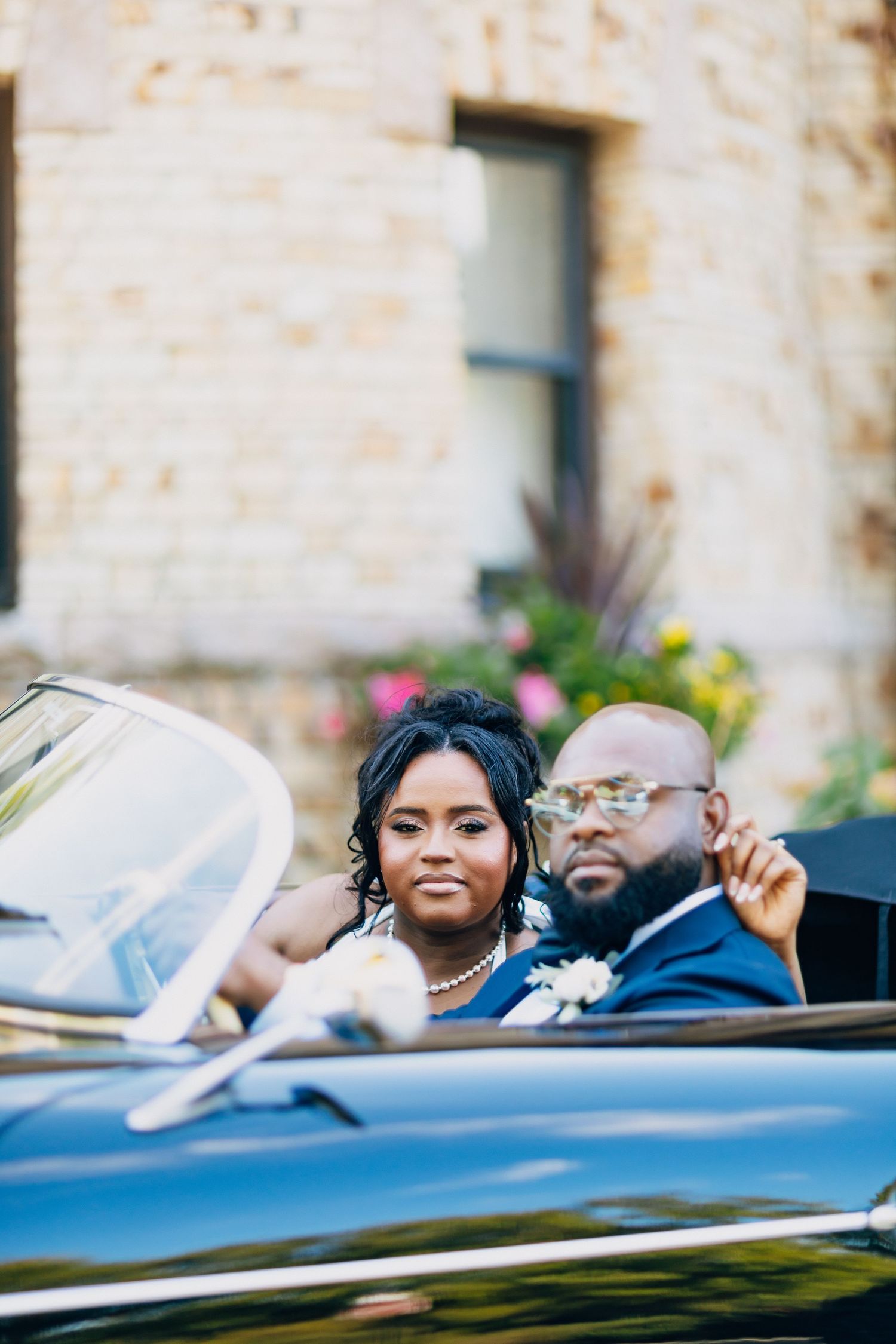 Bride and groom sit in a classic black convertible car outside a stone building wedding venue.