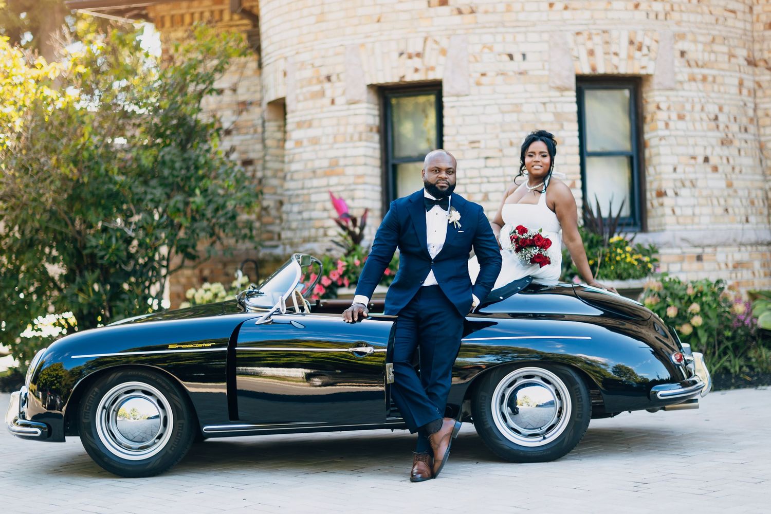 Bride and groom in formal wedding attire posing beside a classic black vintage Porsche convertible outdoors.