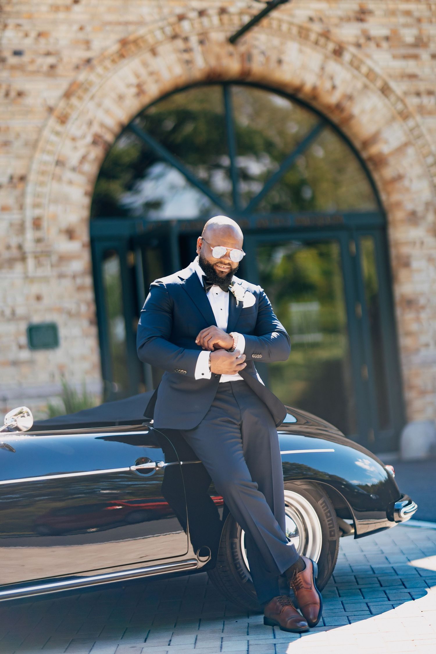 Stylish groom in navy suit leaning against a classic black car outside brick arch building.