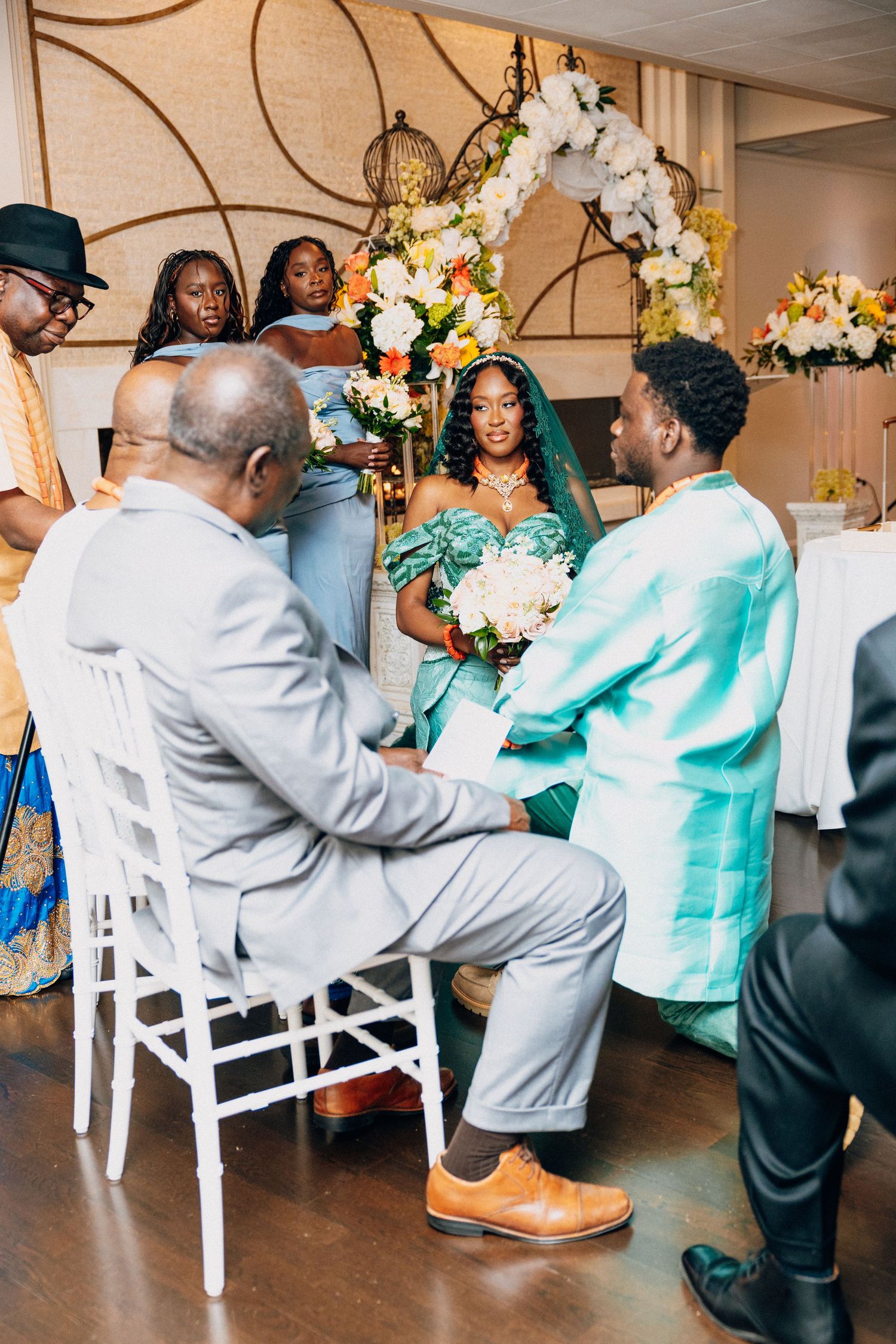 Groom kneels before bride in teal dress during traditional African wedding ceremony.