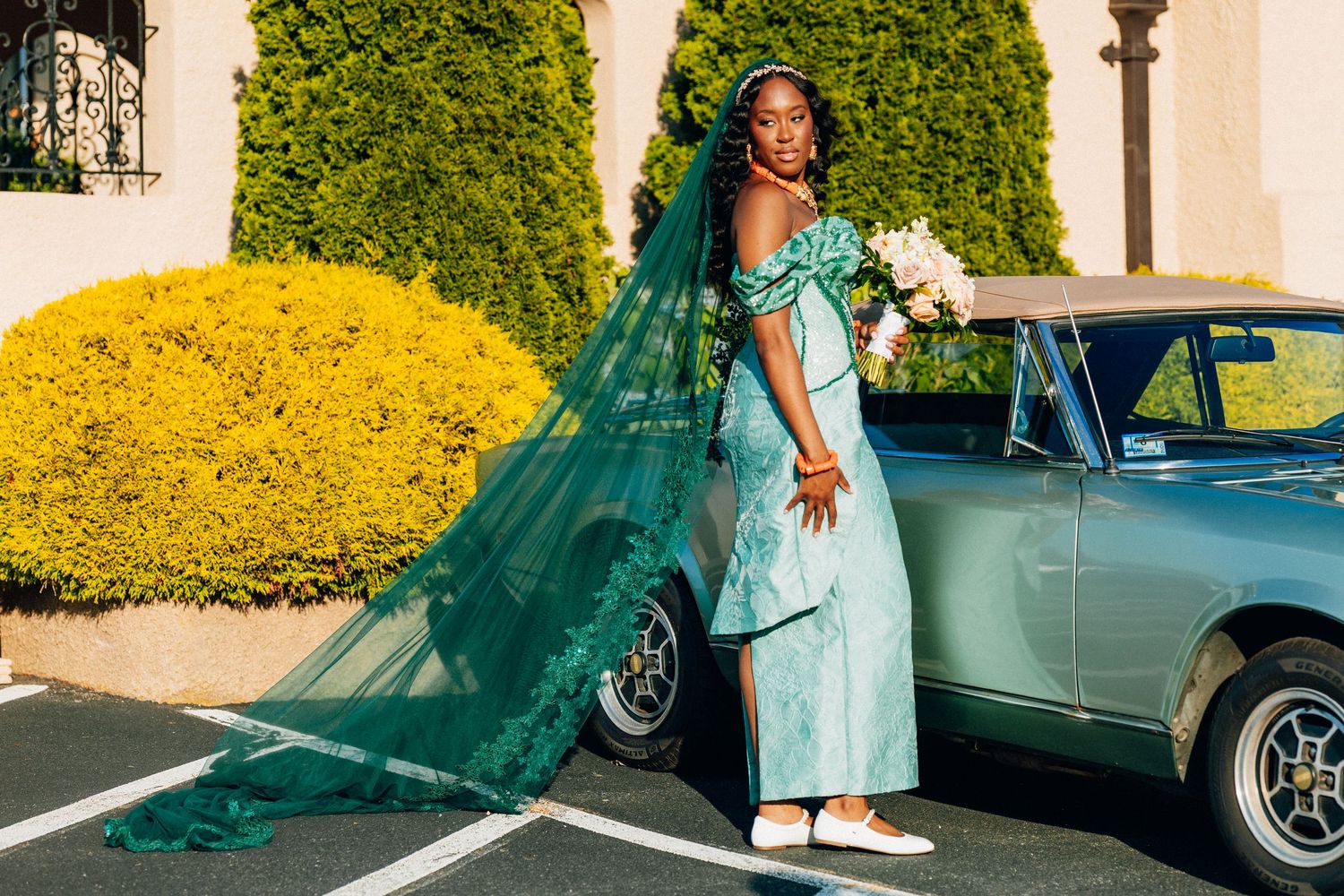 Nigerian bride in mint green gown with long teal veil poses beside vintage green car outdoors.