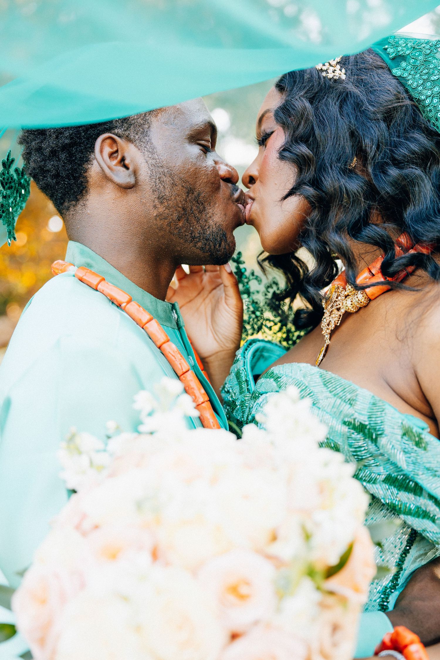 Nigerian bride and groom sharing a kiss under teal fabric with coral beads and bouquet.