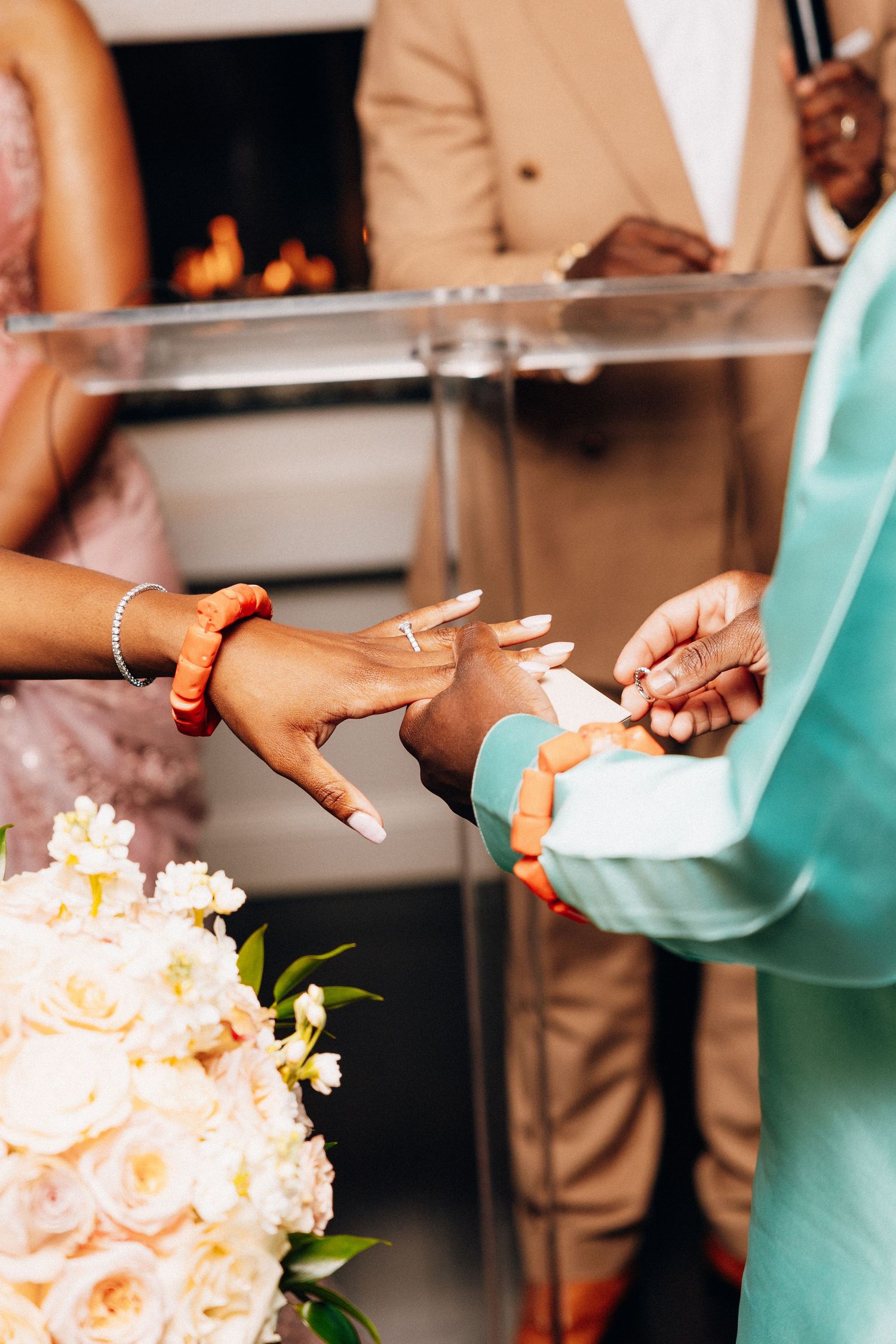 Close-up of a wedding ring exchange ceremony with orange bracelets, white roses bouquet, and a clear acrylic podium.