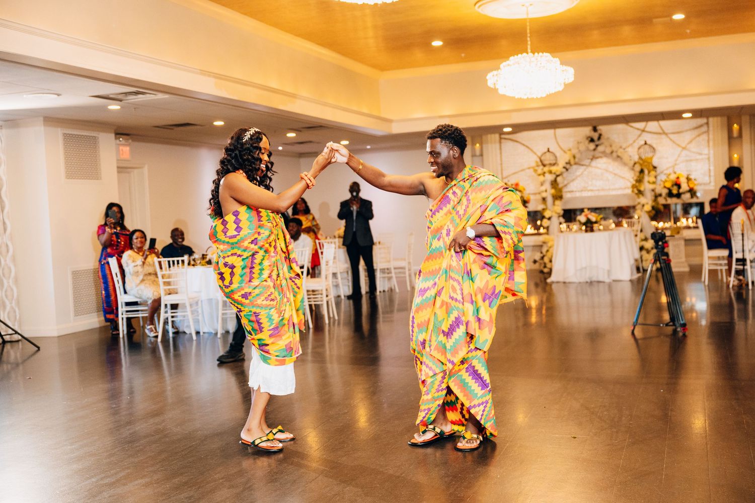 A couple in colorful traditional Kente cloth attire dancing joyfully at their African wedding reception.