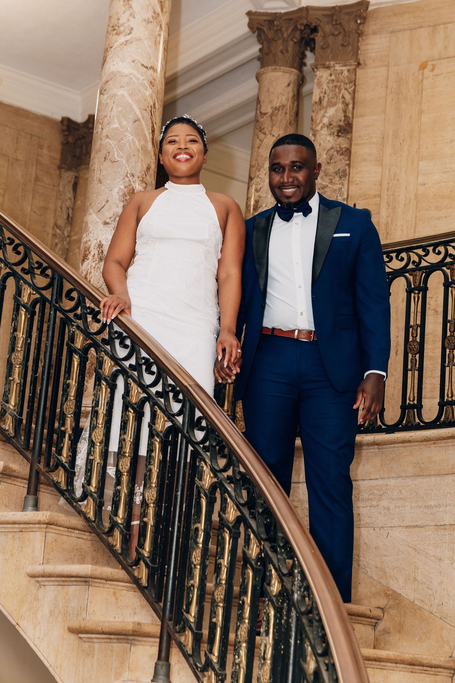 Bride in white halter dress and groom in navy tuxedo hold hands on elegant marble staircase.