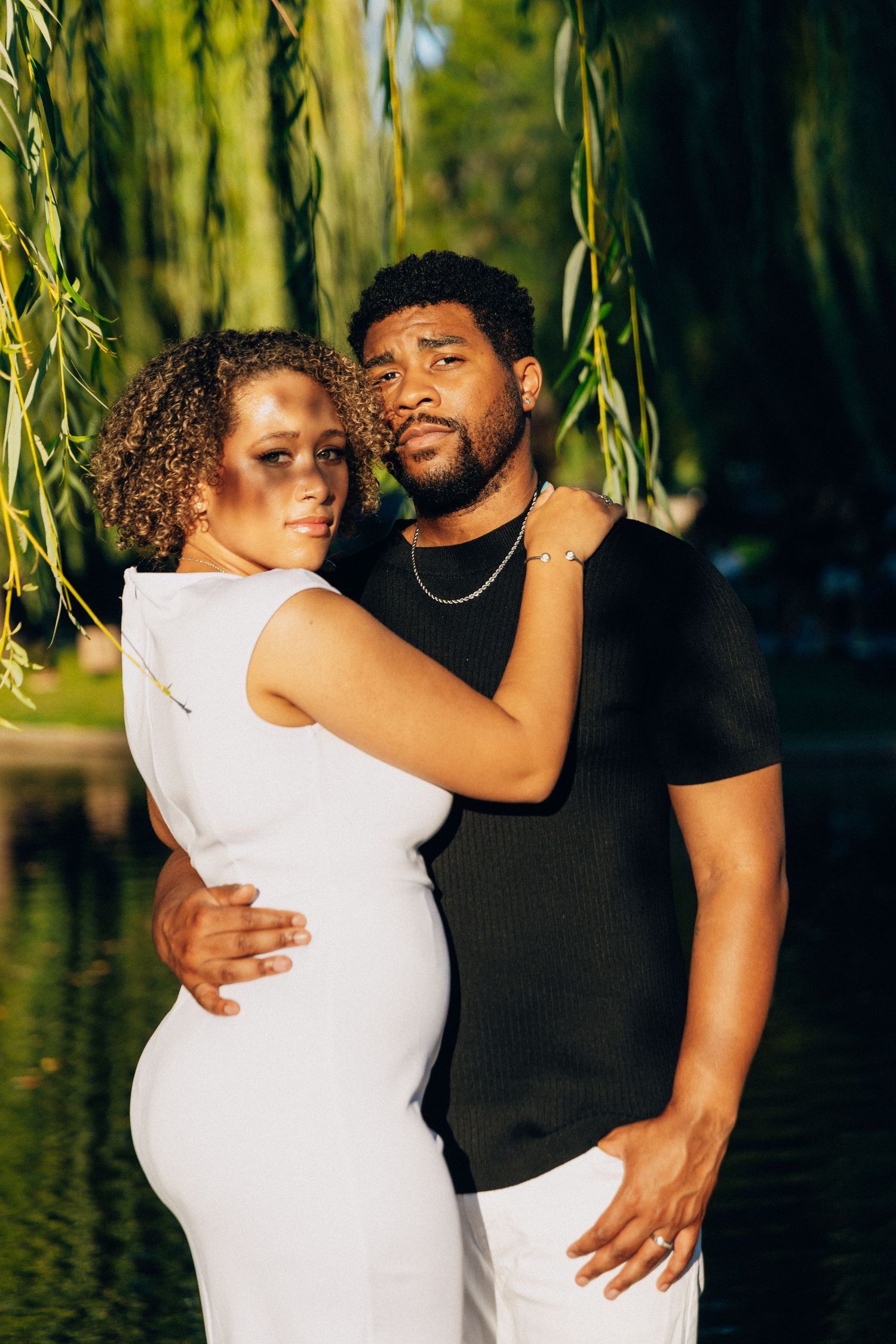 A couple poses closely together near a pond under willow tree branches, woman in white dress, man in black shirt.