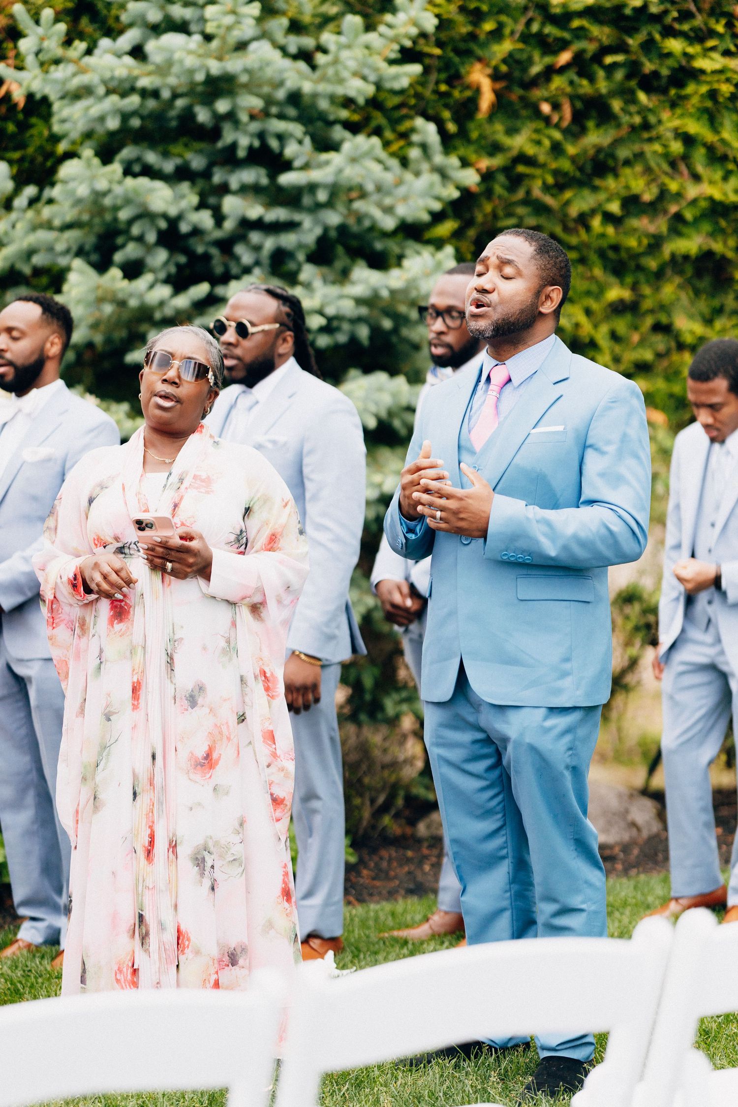 Wedding guests in blue suits and floral dress sing during outdoor garden ceremony.