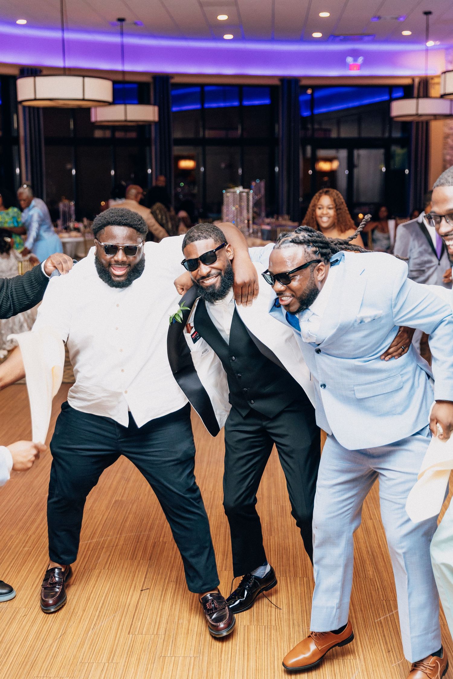 Groomsmen wearing sunglasses dance and celebrate joyfully on the dance floor at a wedding reception.