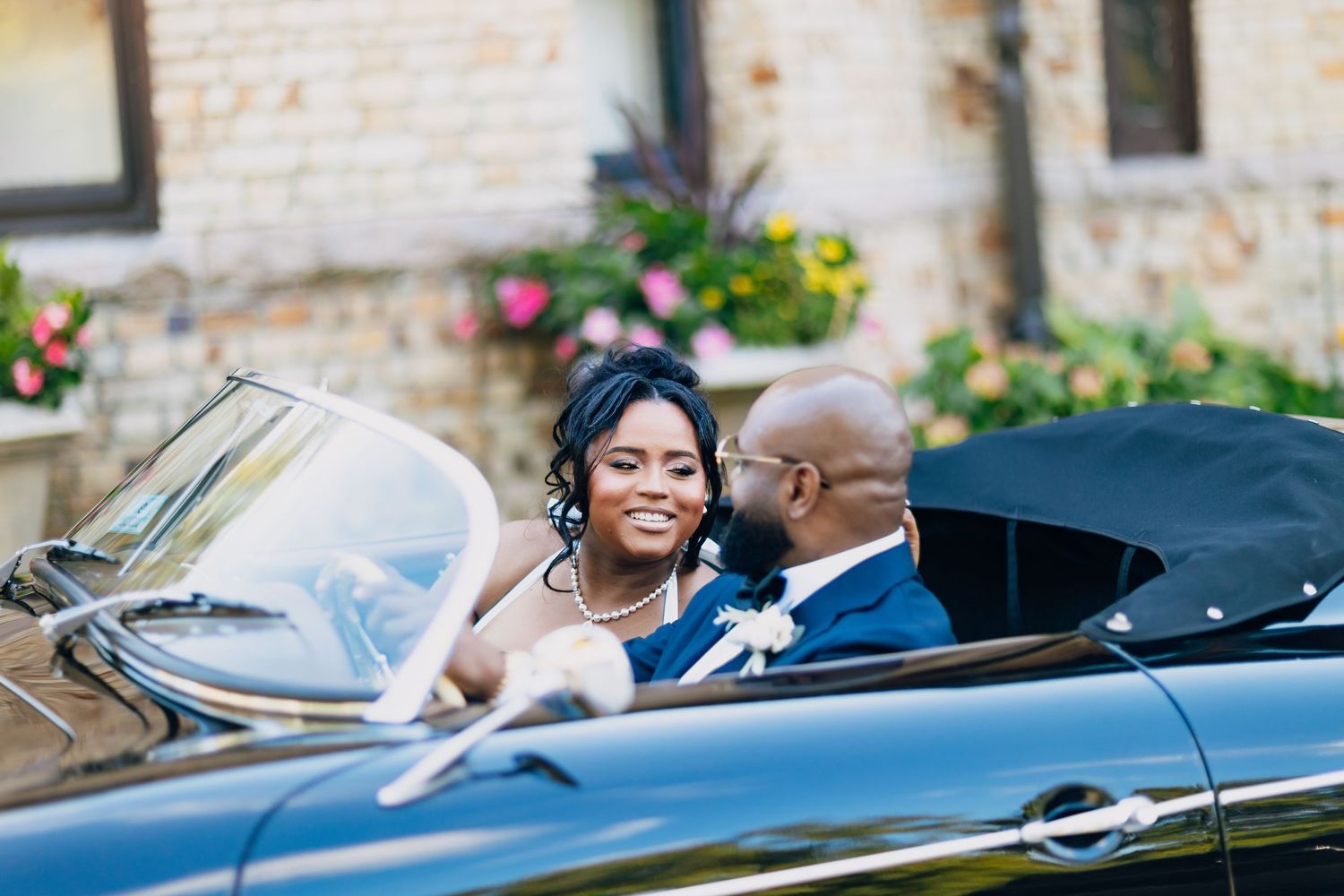 Smiling bride looks at groom while seated together in a vintage black convertible wedding car.