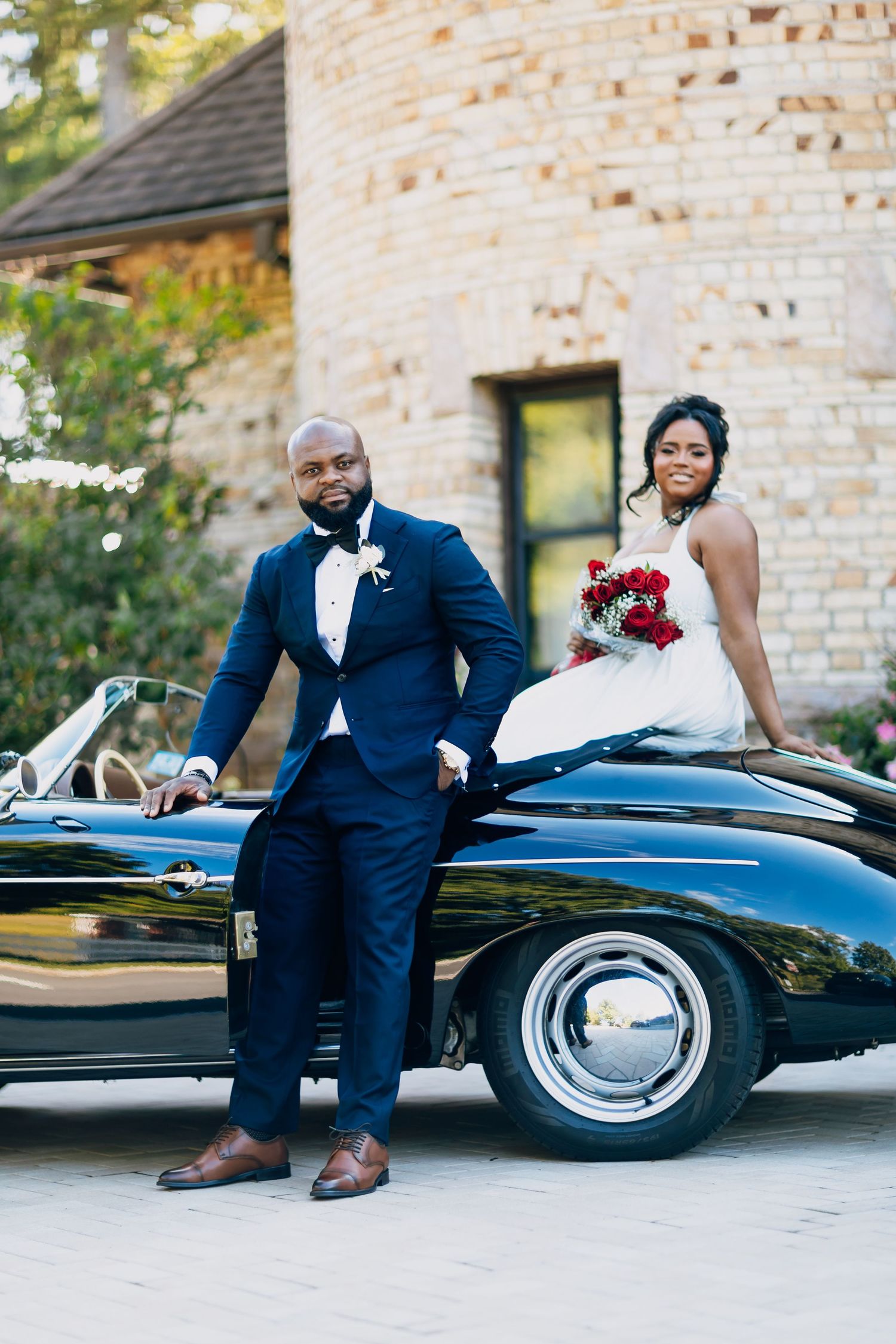 Groom leaning on a classic black Porsche as bride sits on the car holding a red bouquet.