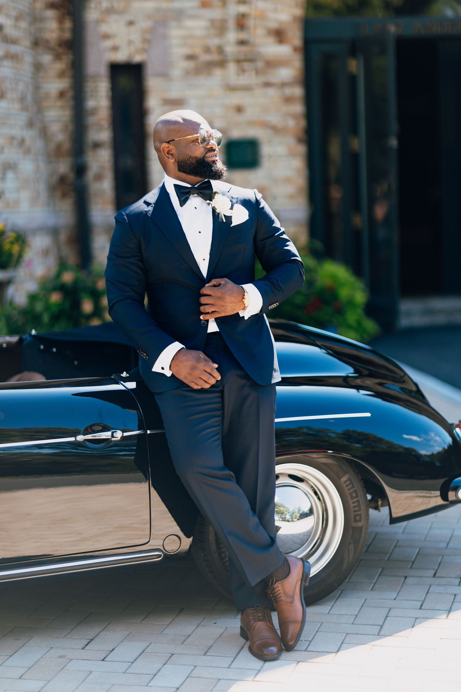 Dapper groom in navy tuxedo with bow tie leans against a classic black vintage car outdoors.