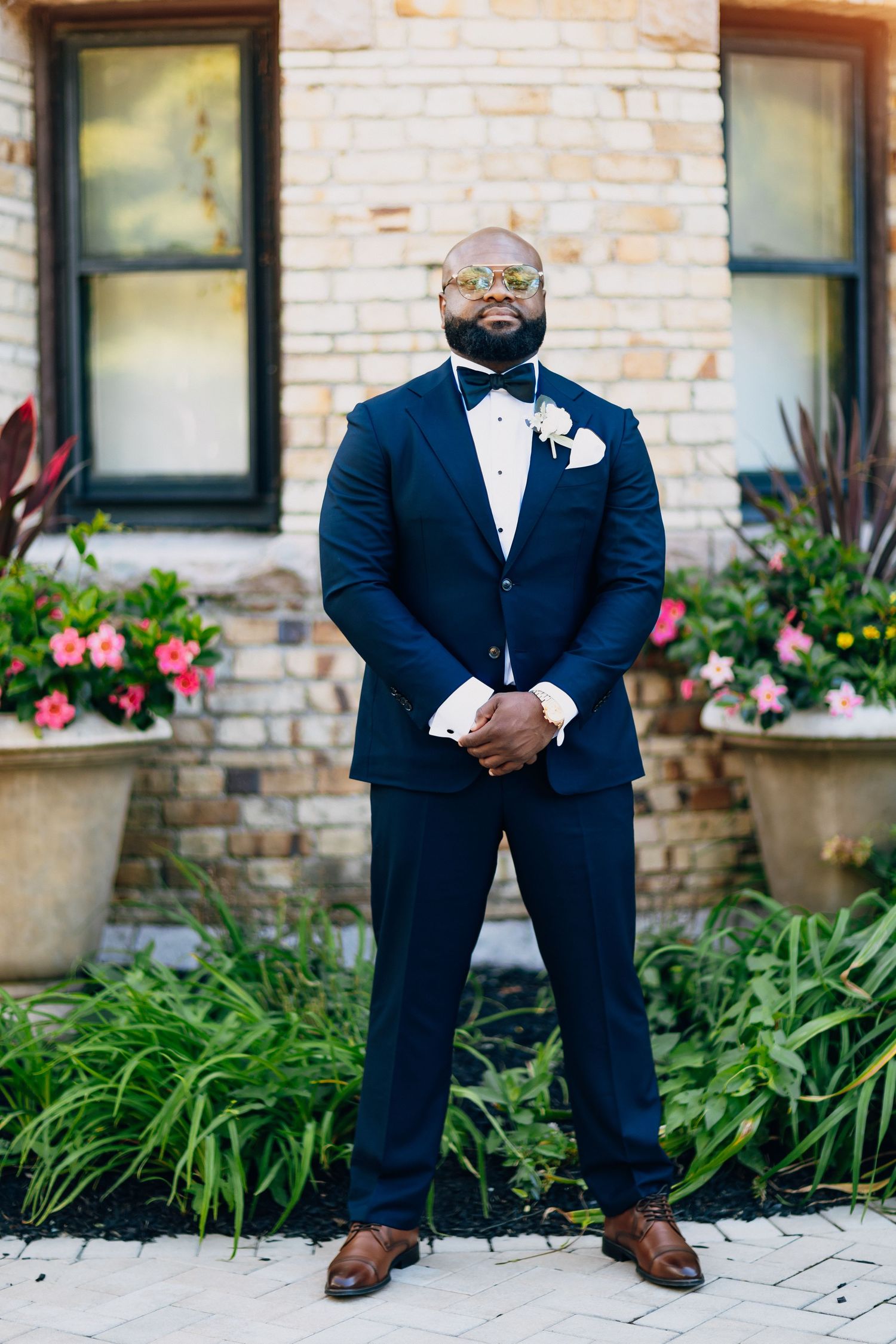Groom in navy blue tuxedo with bow tie and boutonniere stands confidently before brick building with flowers.