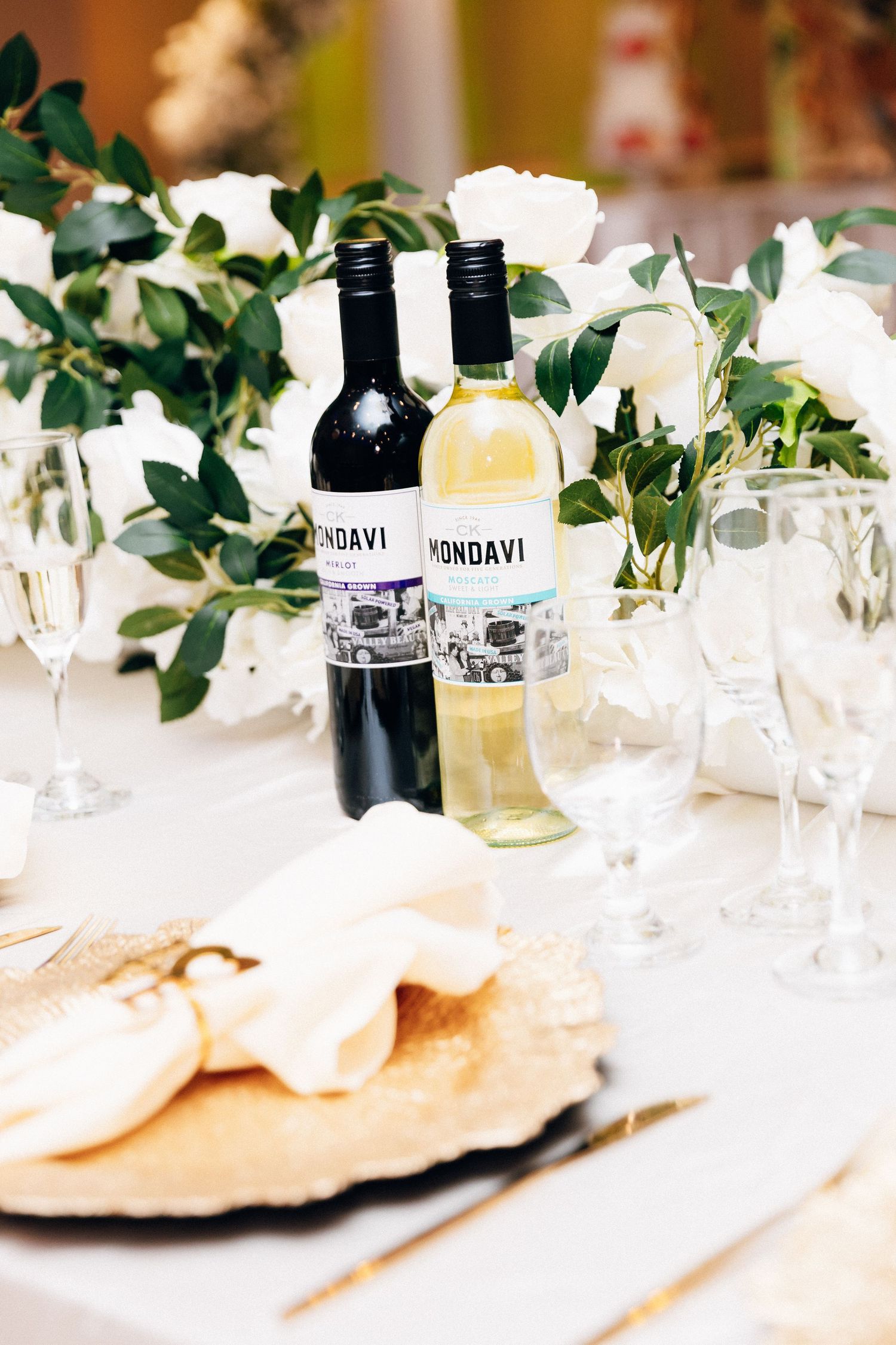 Mondavi red and white wine bottles on elegant wedding table with white roses, greenery, and champagne flutes.