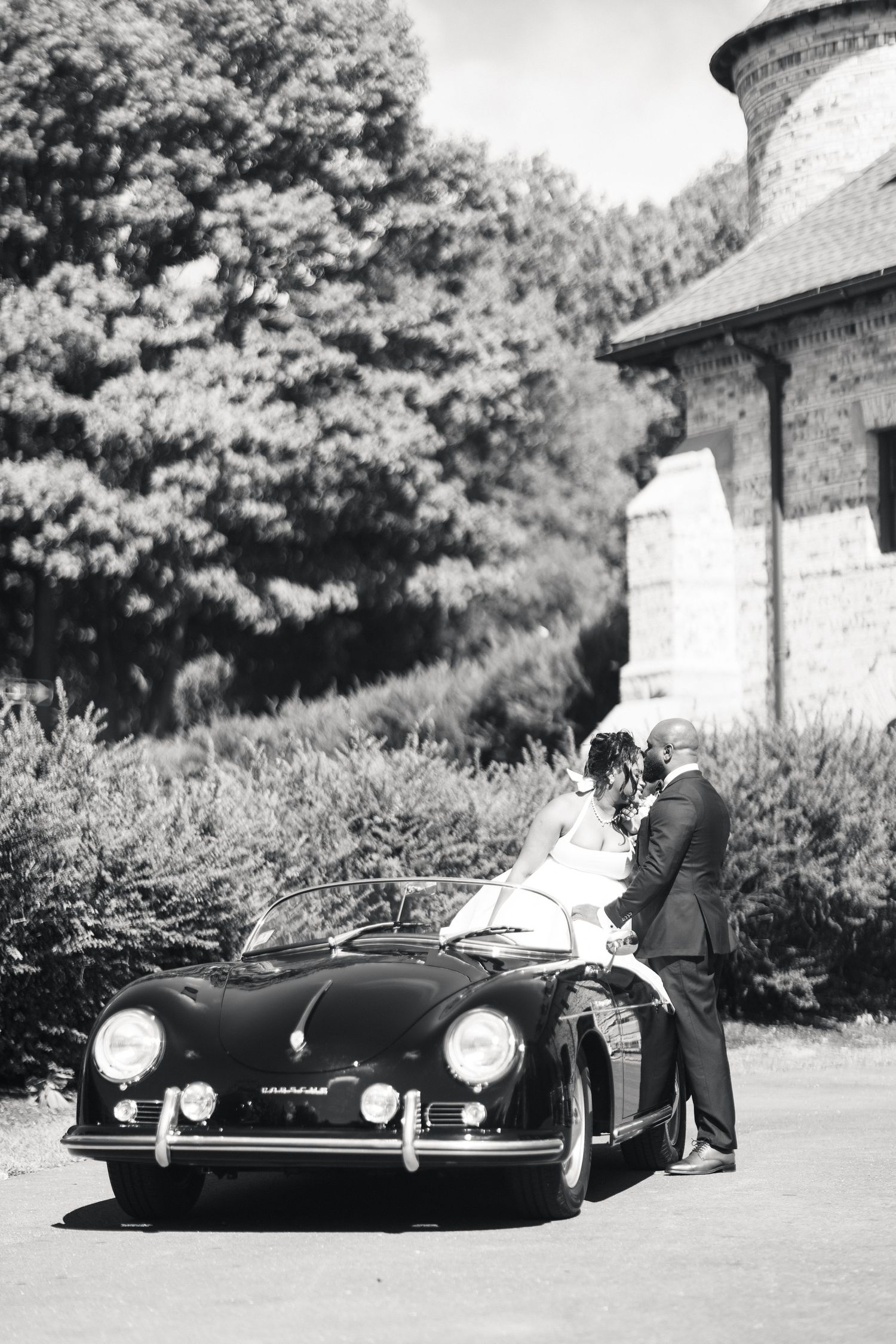 Black and white photo of newlyweds standing beside a classic Porsche 356 at brick venue.
