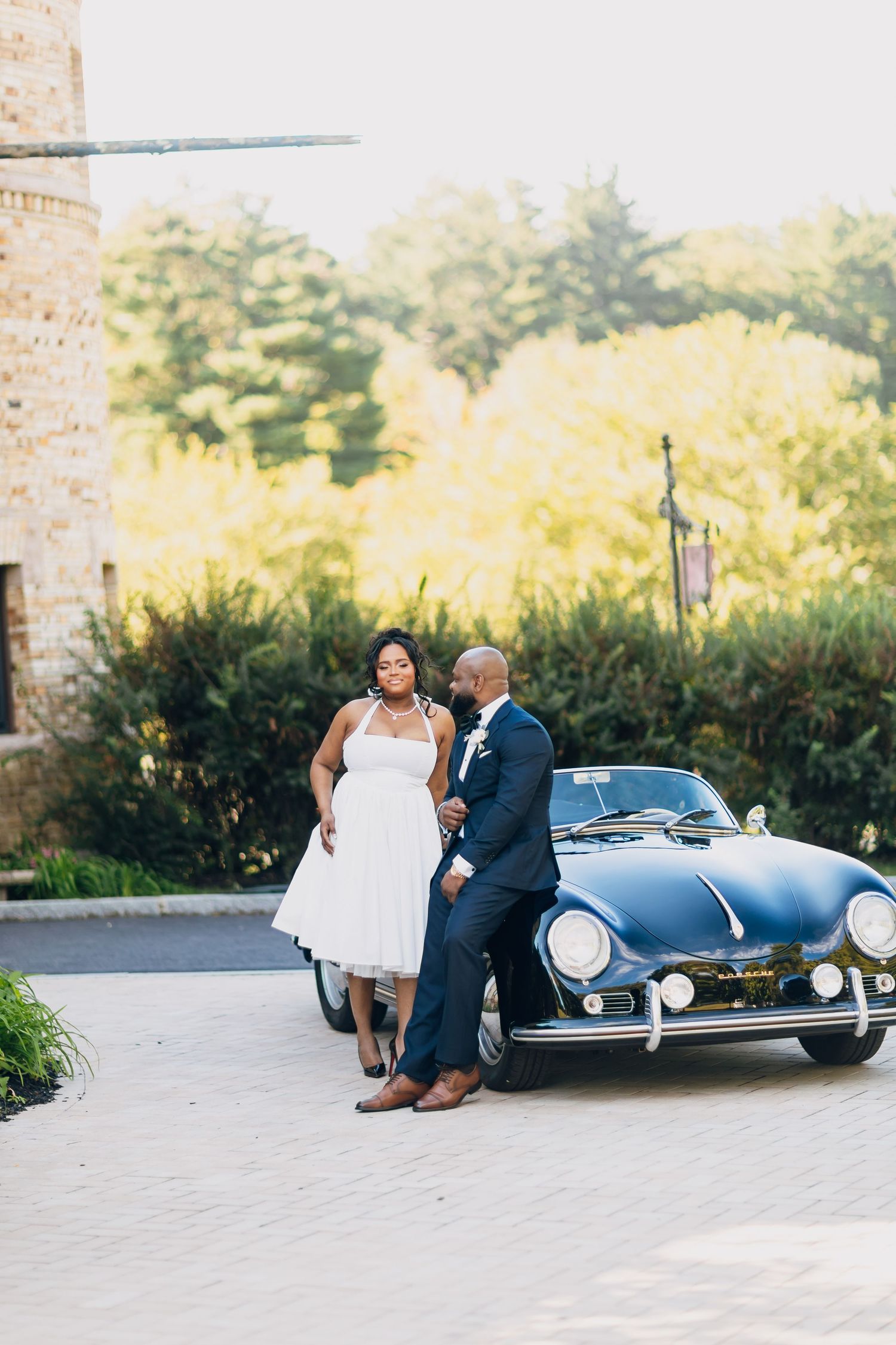 A bride and groom pose beside a classic vintage navy Porsche on their wedding day outdoors.