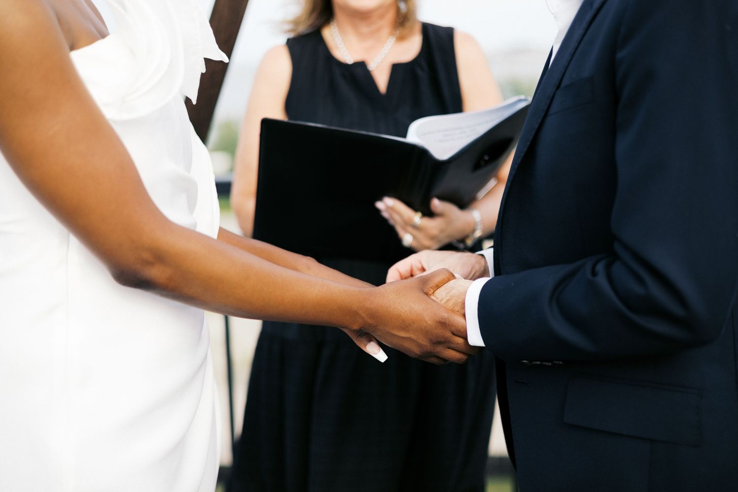 Bride and groom holding hands during wedding ceremony with officiant in black holding book.