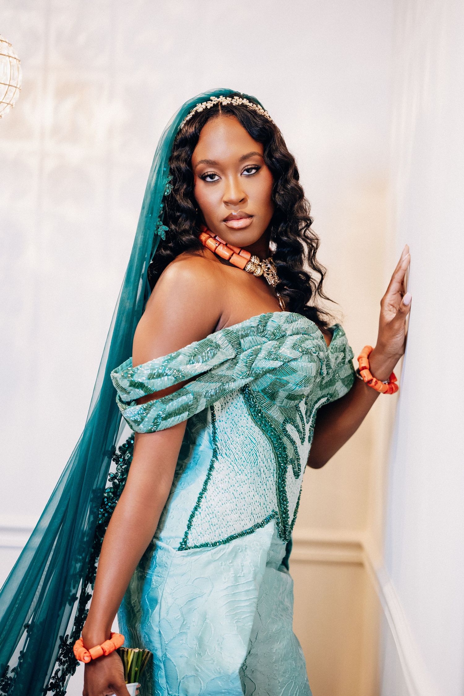 Black bride in teal off-shoulder Nigerian traditional attire with coral beads and floral headpiece.
