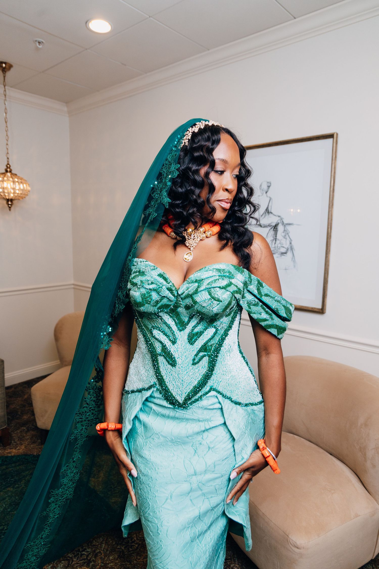 Bride wearing a stunning teal green African wedding gown with embroidery, veil, and orange beaded jewelry.
