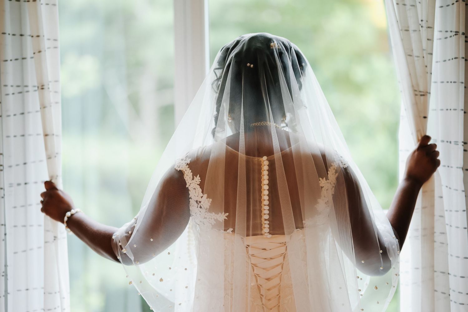 Black bride in lace wedding dress and veil stands at window, viewed from behind.
