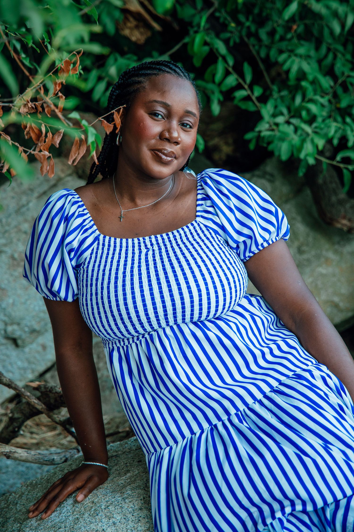 A young Black woman in a blue and white striped dress sits outdoors smiling, surrounded by lush green foliage.