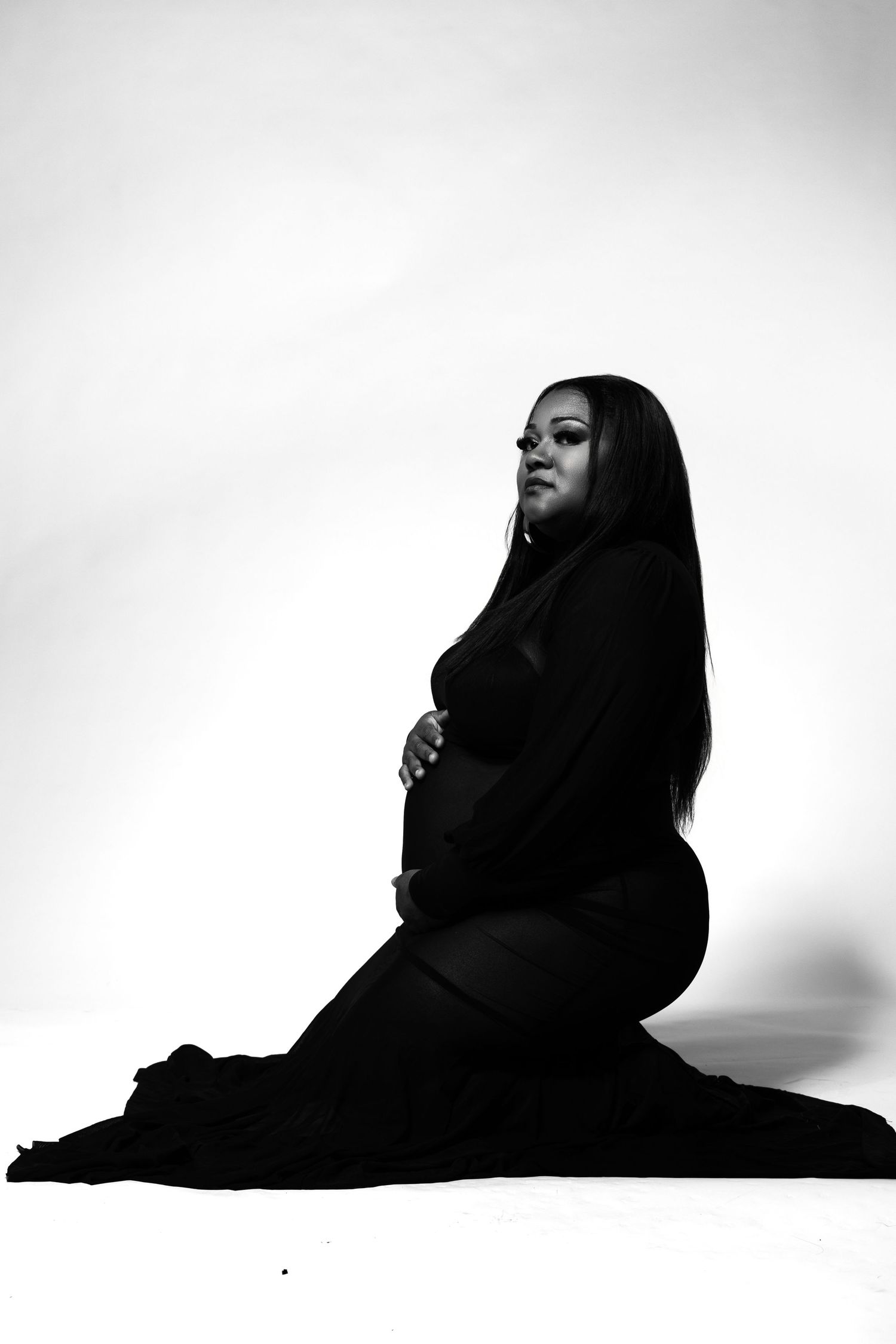 Black and white maternity photo of a pregnant woman in a black dress kneeling gracefully in a studio setting.