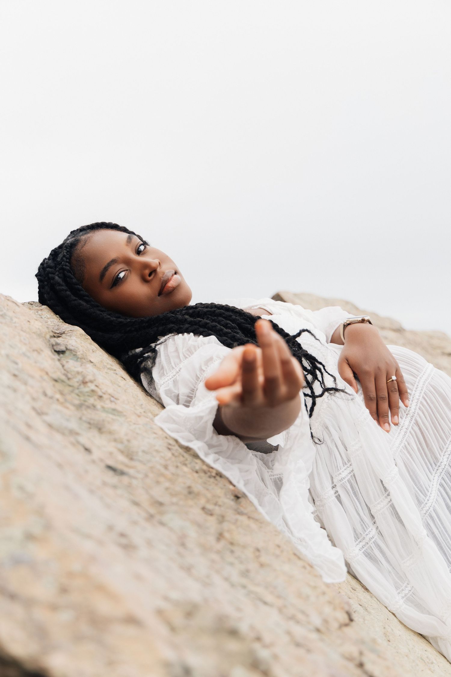 A Black woman in a white dress reclines on rocks, reaching toward the camera with long braided hair flowing around her.