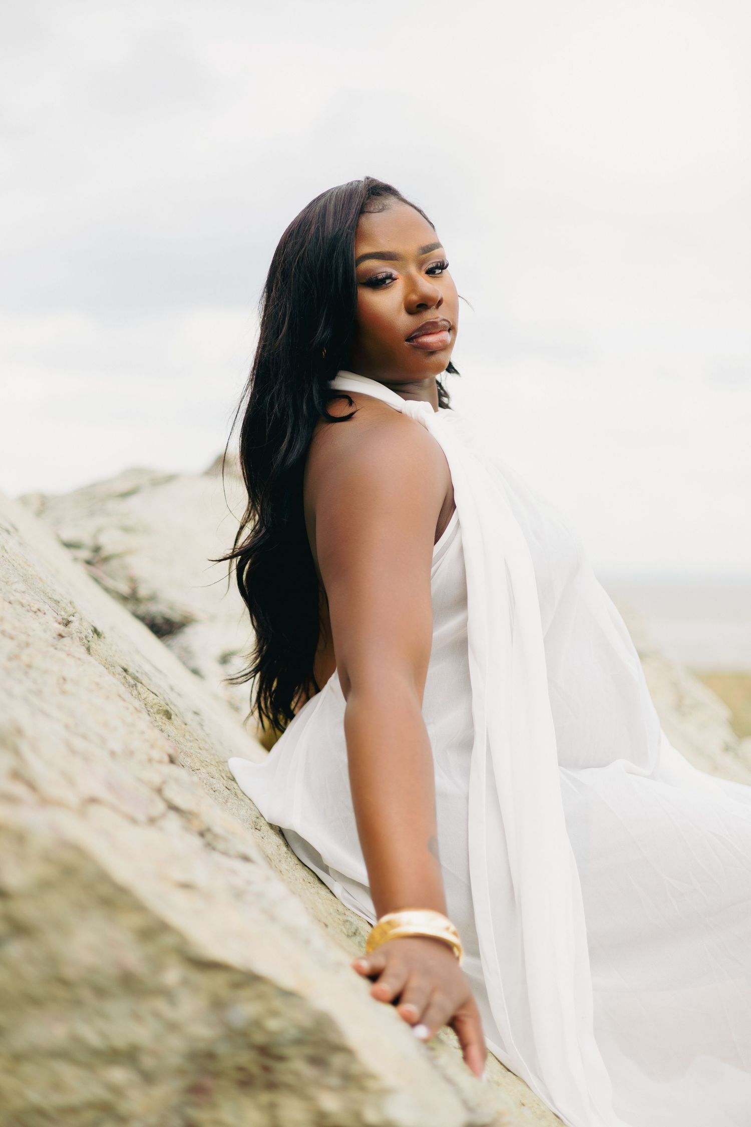 A Black woman in a flowing white halter dress and gold bracelet poses gracefully on rocky terrain with an overcast sky.
