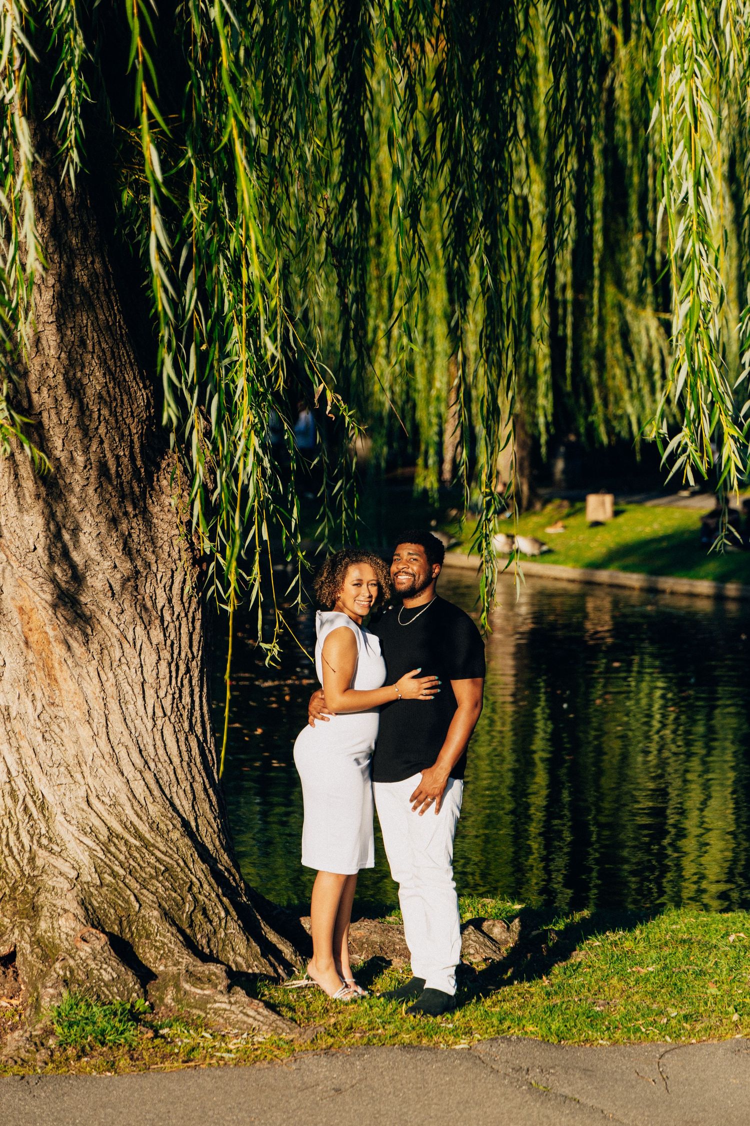 A smiling couple poses under a weeping willow tree by a tranquil pond, dressed in black and white.