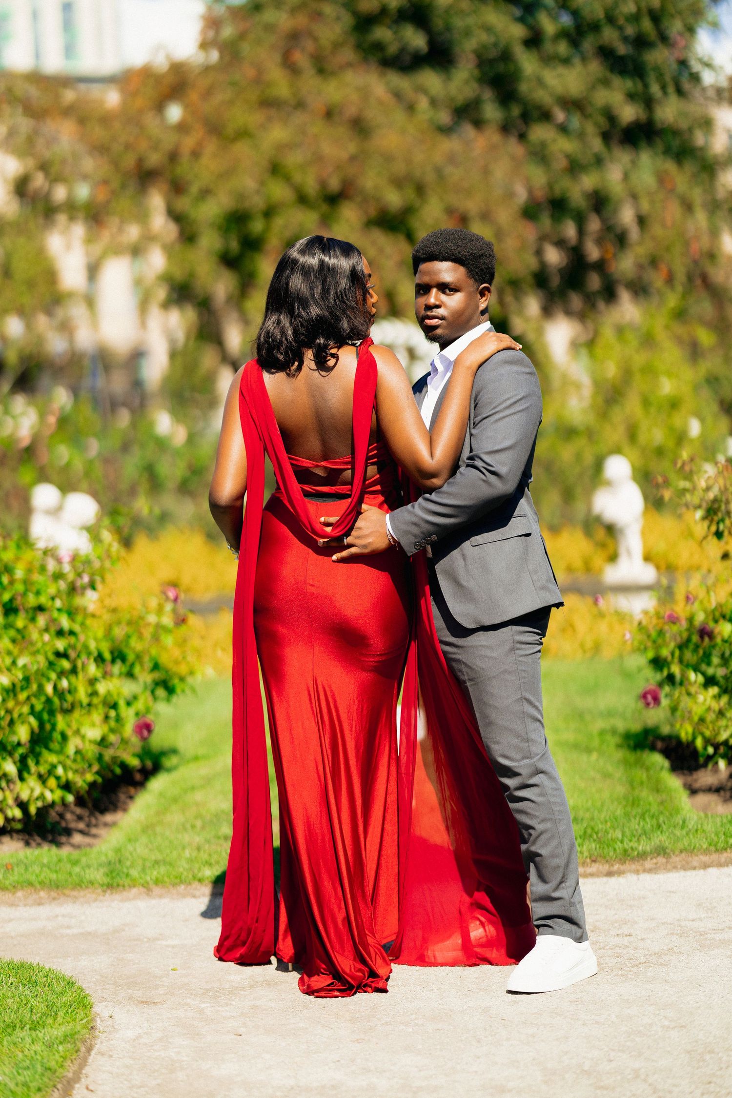 A couple in formal attire poses in a garden, woman in red gown, man in gray suit, surrounded by lush greenery.