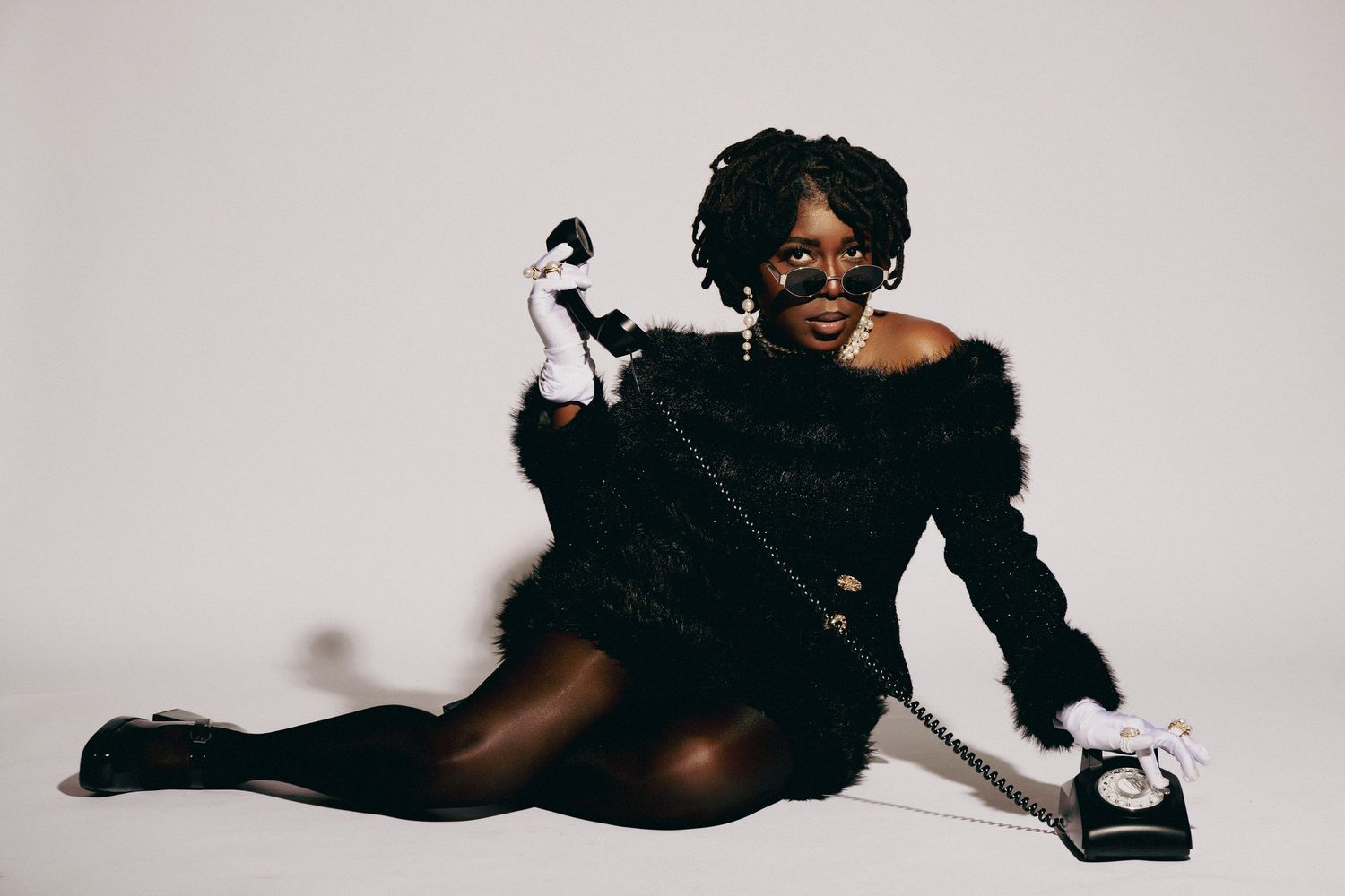 A stylish person in black fur jacket poses on floor holding vintage telephone handset with sunglasses on white background.
