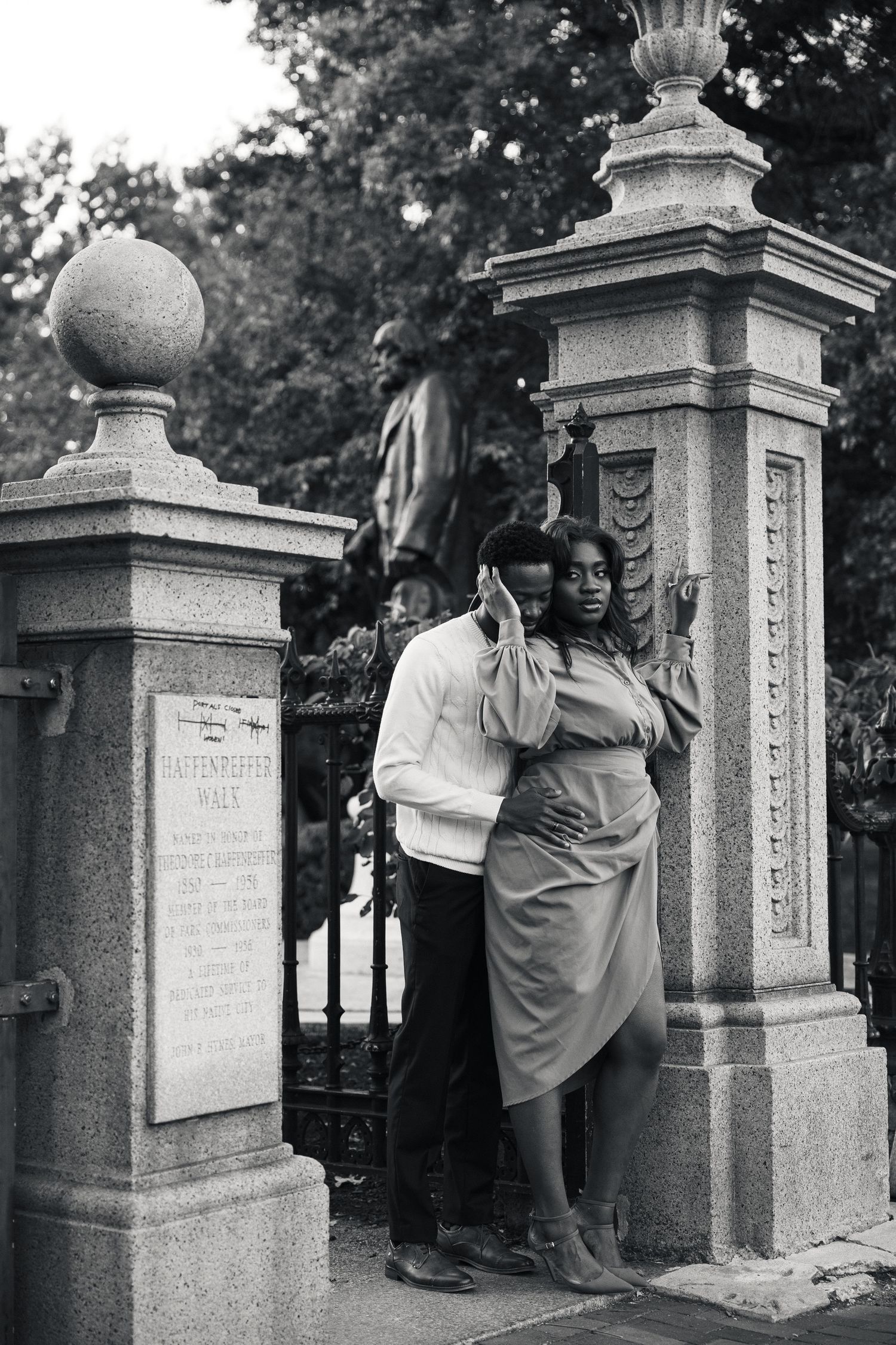 Black and white photo of a couple posing romantically at ornate stone gate pillars in a park setting.