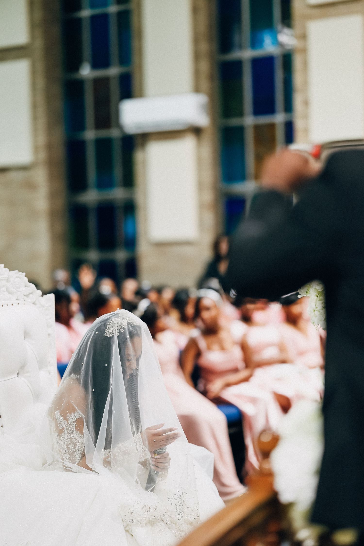Bride in white lace veil seated at church wedding ceremony with bridesmaids in pink dresses in background.