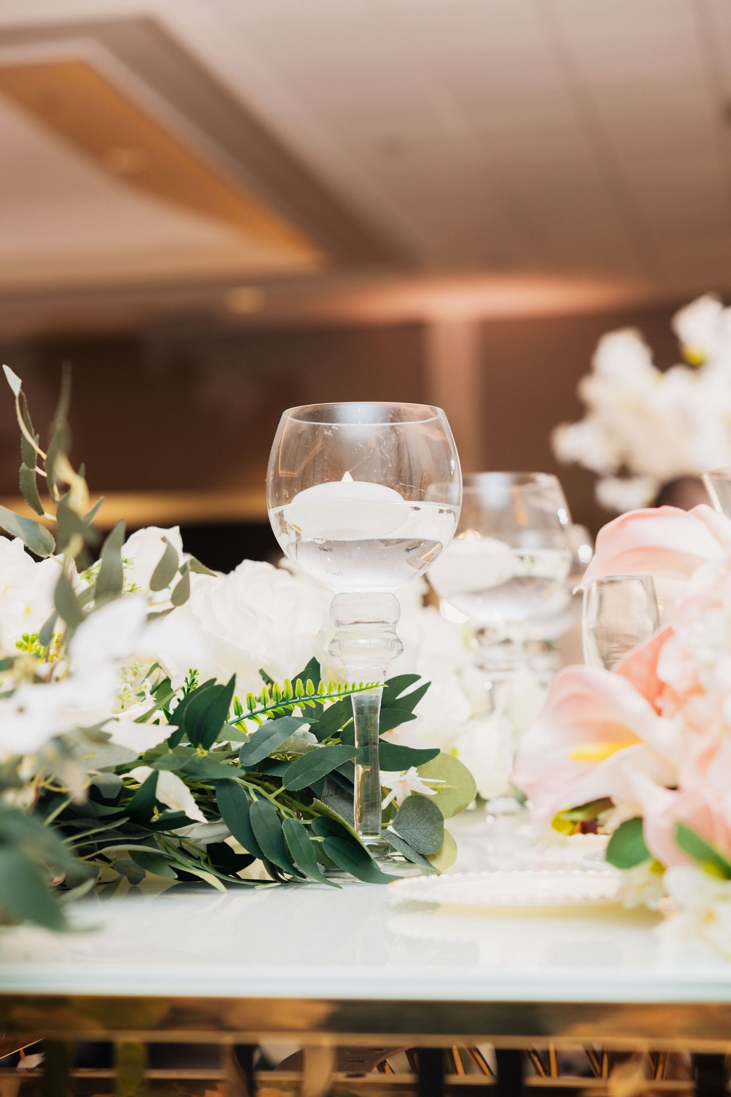 Elegant wedding table with white roses, greenery, and a glass candle holder centerpiece in a softly lit reception hall.