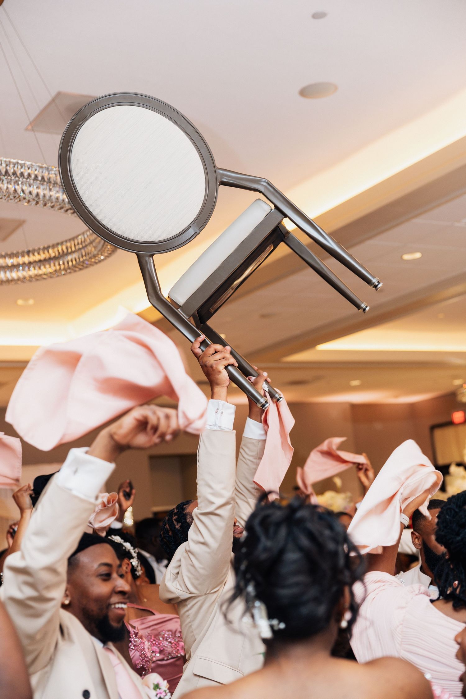 Energetic wedding guests lift chair in celebration during lively reception dance with pink napkins waving.