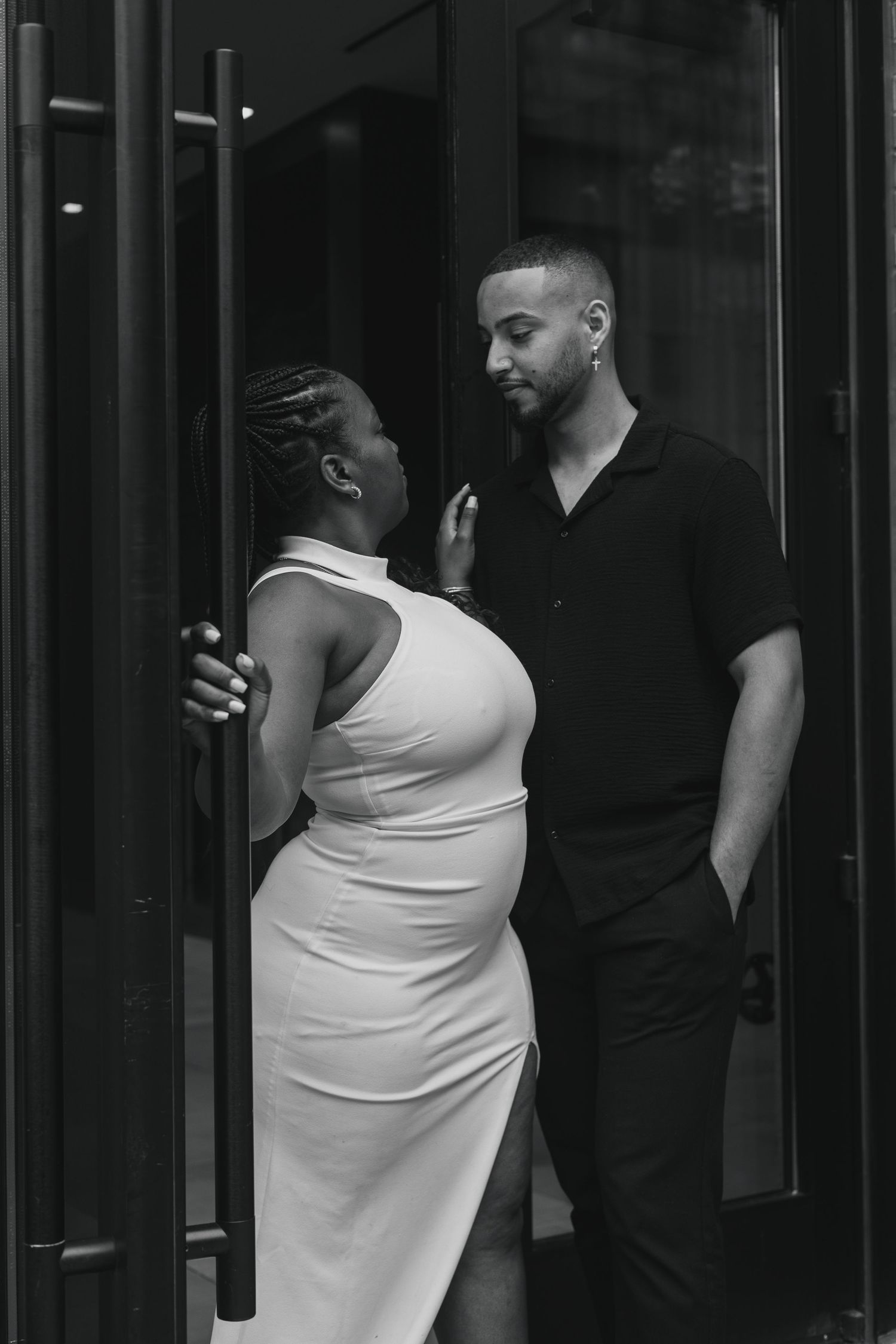 Black and white photo of a pregnant woman in white dress and man in black outfit sharing an intimate moment in a doorway.