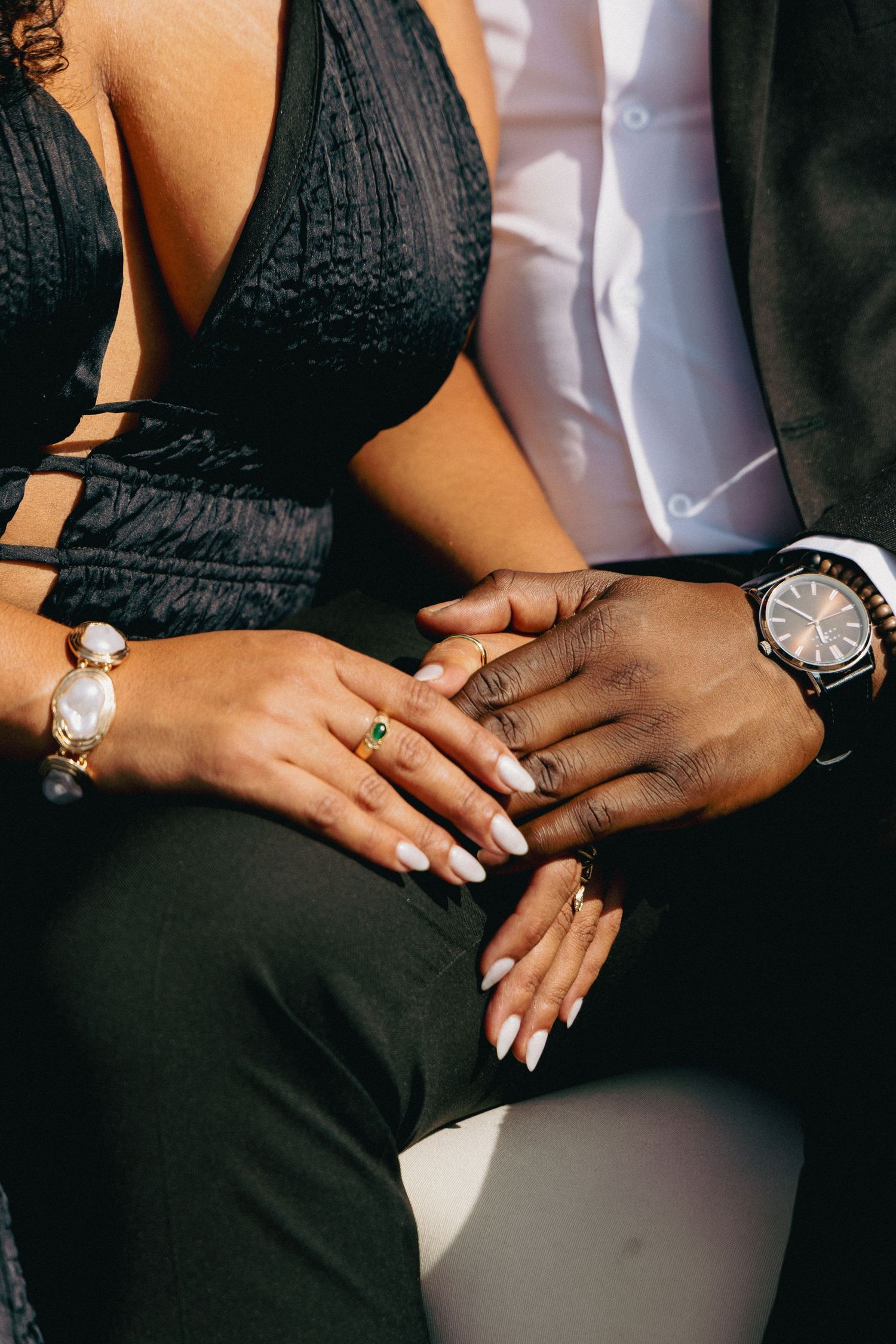 Close-up of an interracial couple holding hands, showcasing elegant rings, bracelets, and a watch while dressed in formal attire.