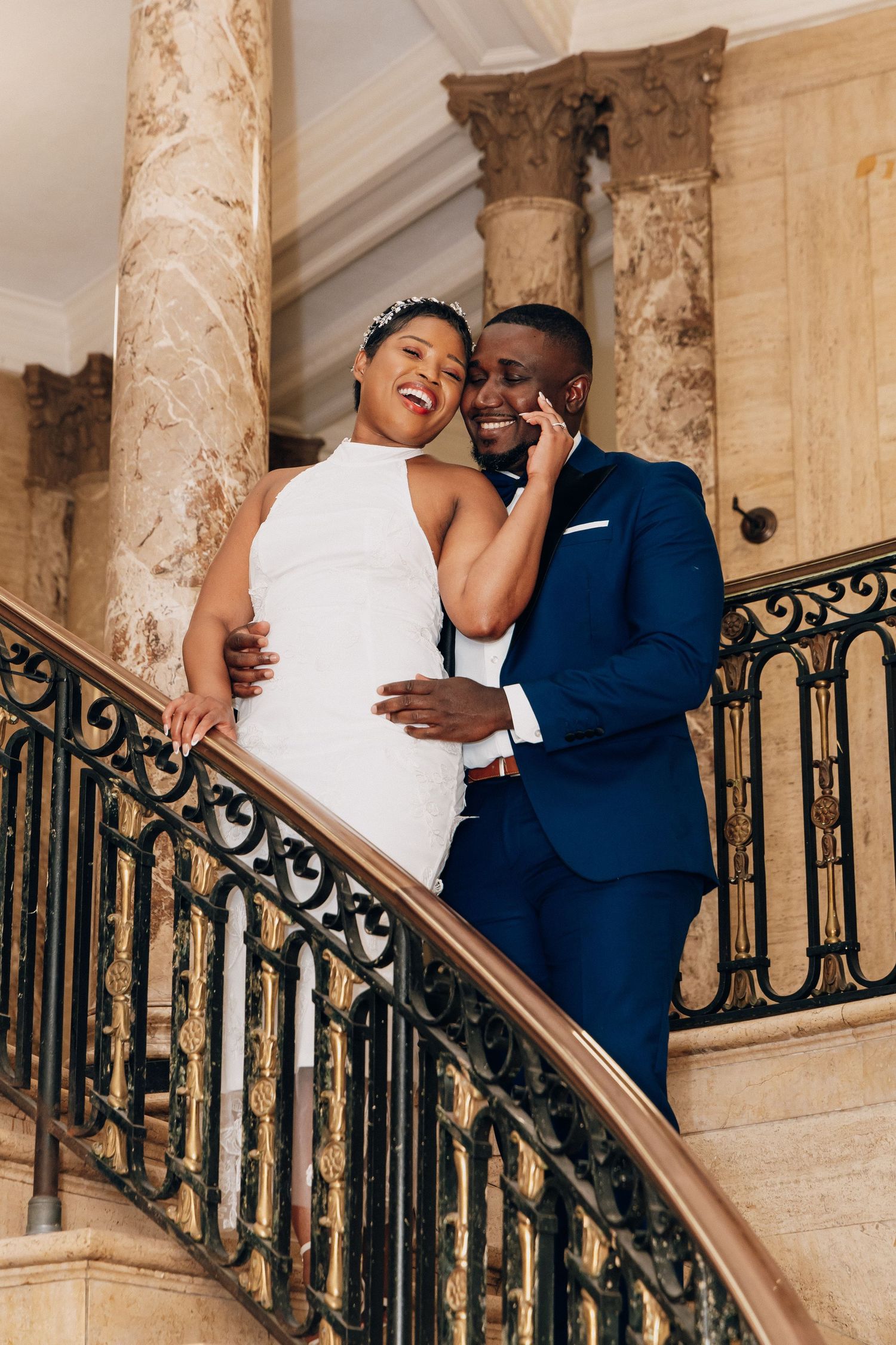 Bride and groom embrace on ornate marble staircase, she in white halter gown, he in navy suit.