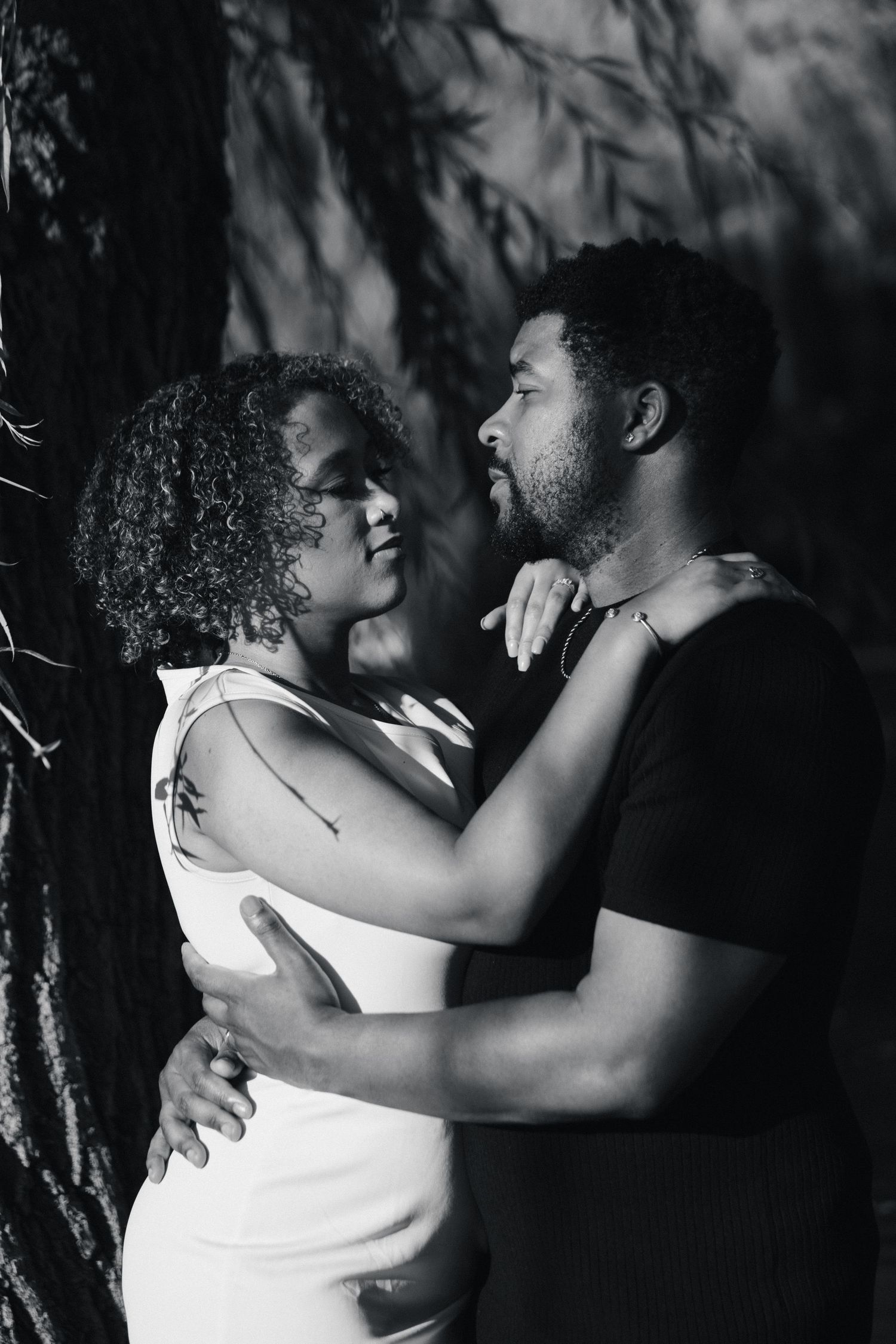 Black and white photo of a romantic couple embracing tenderly near a tree, woman in white dress touching mans face.