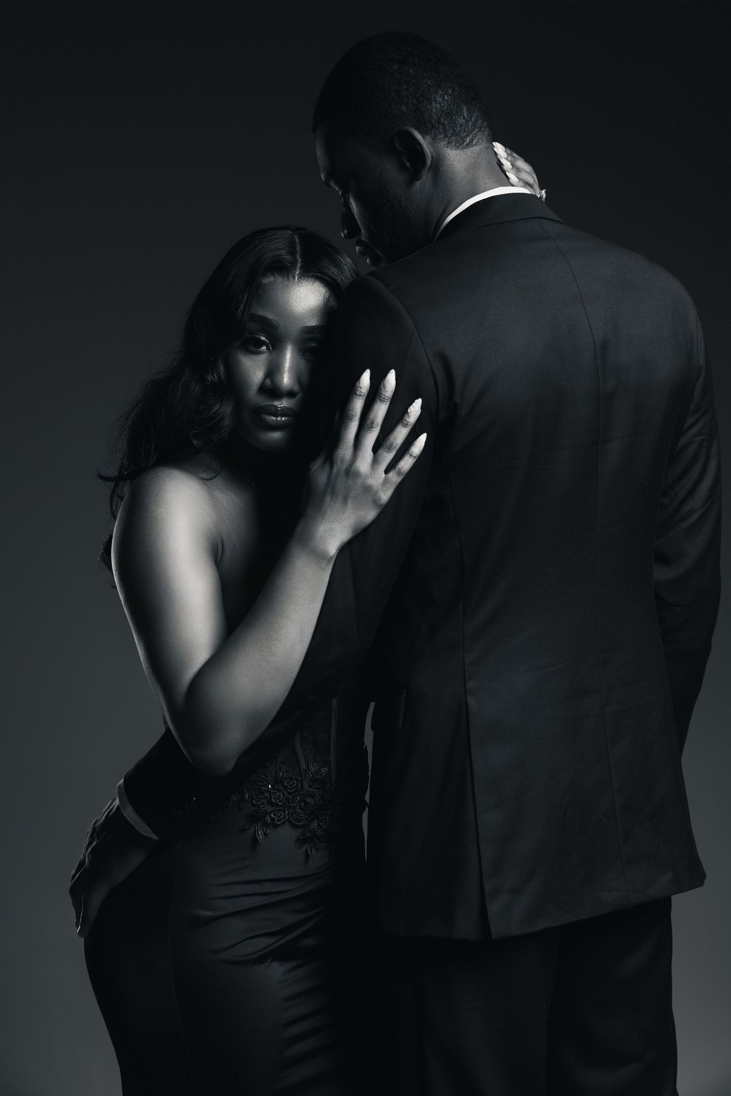 Elegant couple in black and white, woman in gown leans on man in suit in a dramatic studio portrait.