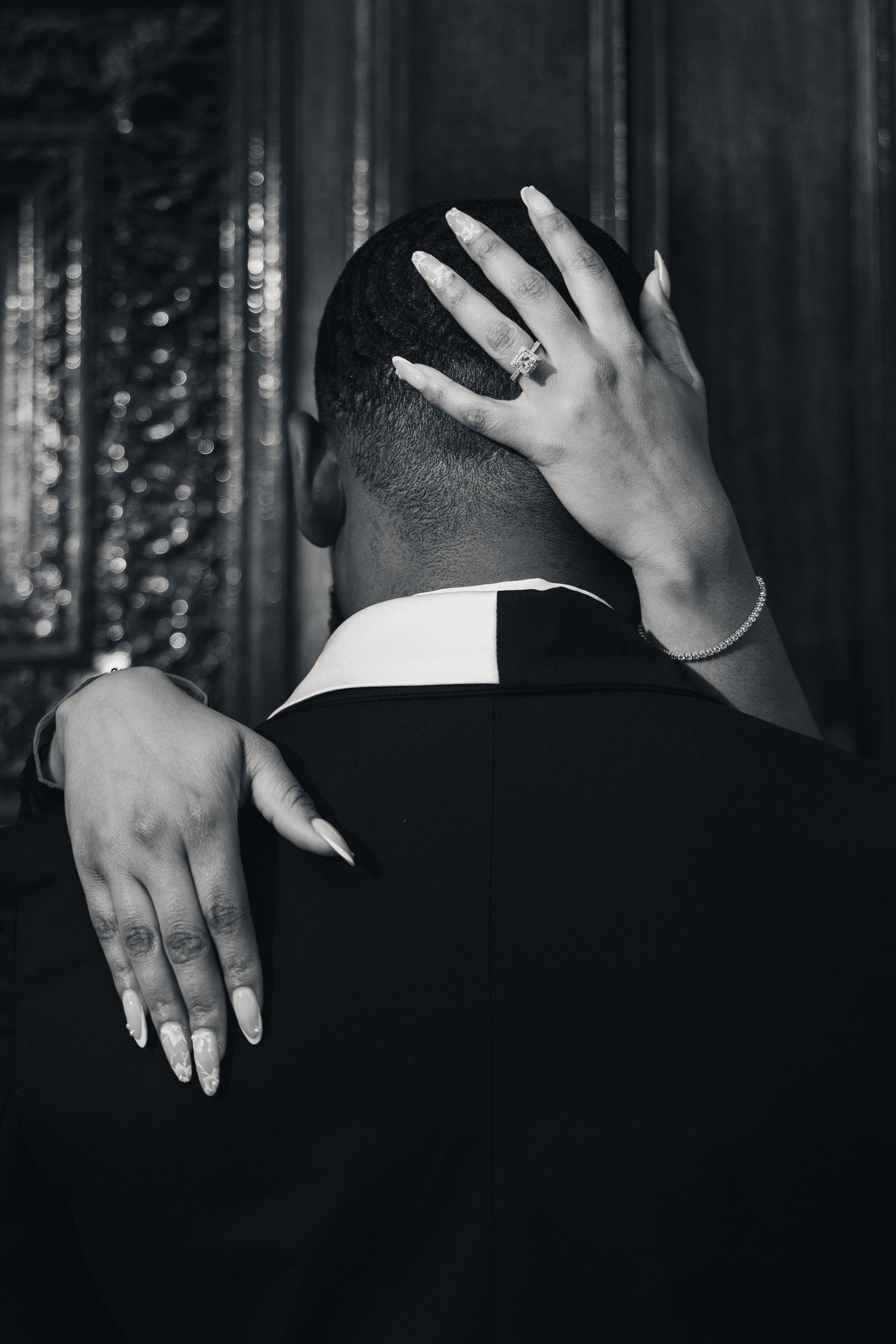 Black and white photo of a couple embracing, woman's hands with ring and bracelet resting on man's head and shoulder.