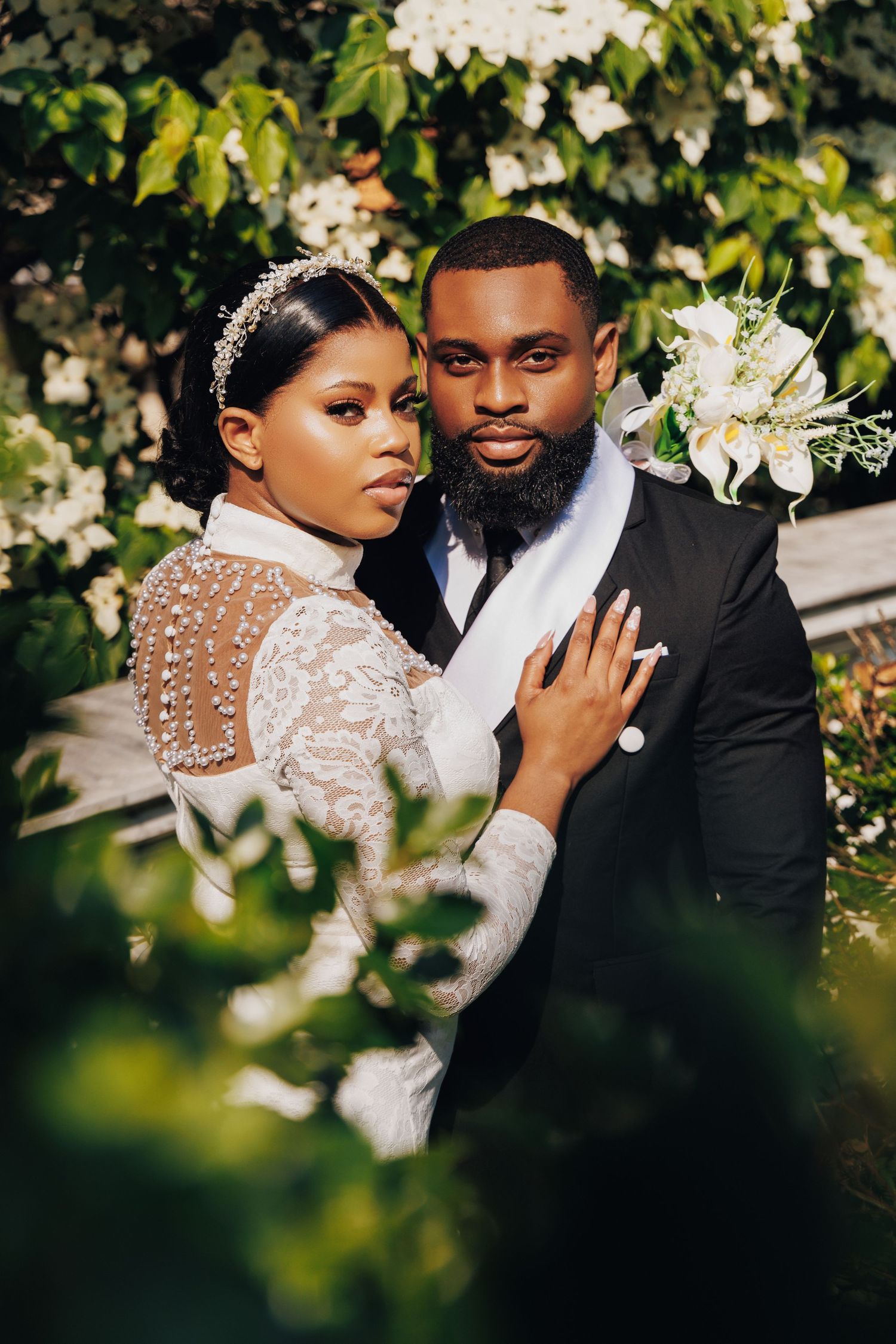 A Black bride and groom pose elegantly among white flowering bushes, she in lace and he in a black suit.