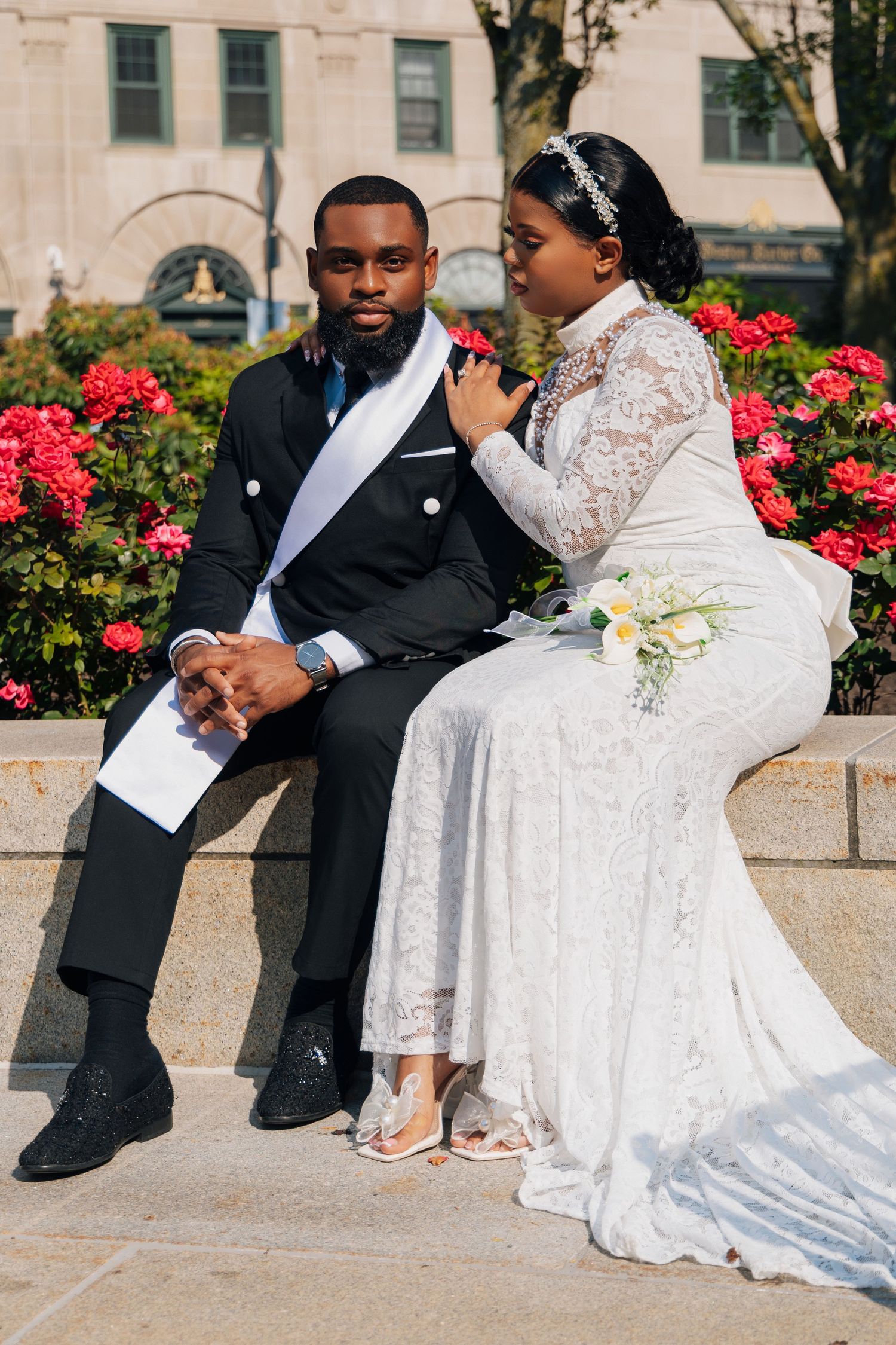 A newlywed couple in wedding attire sits on a stone ledge surrounded by red flowers, sharing a tender moment.