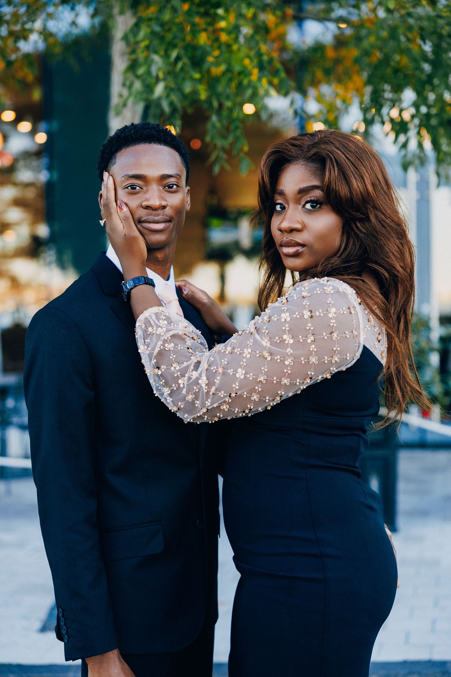 Elegant Black couple posing outdoors, woman in sequined top touching mans face, both wearing formal attire.