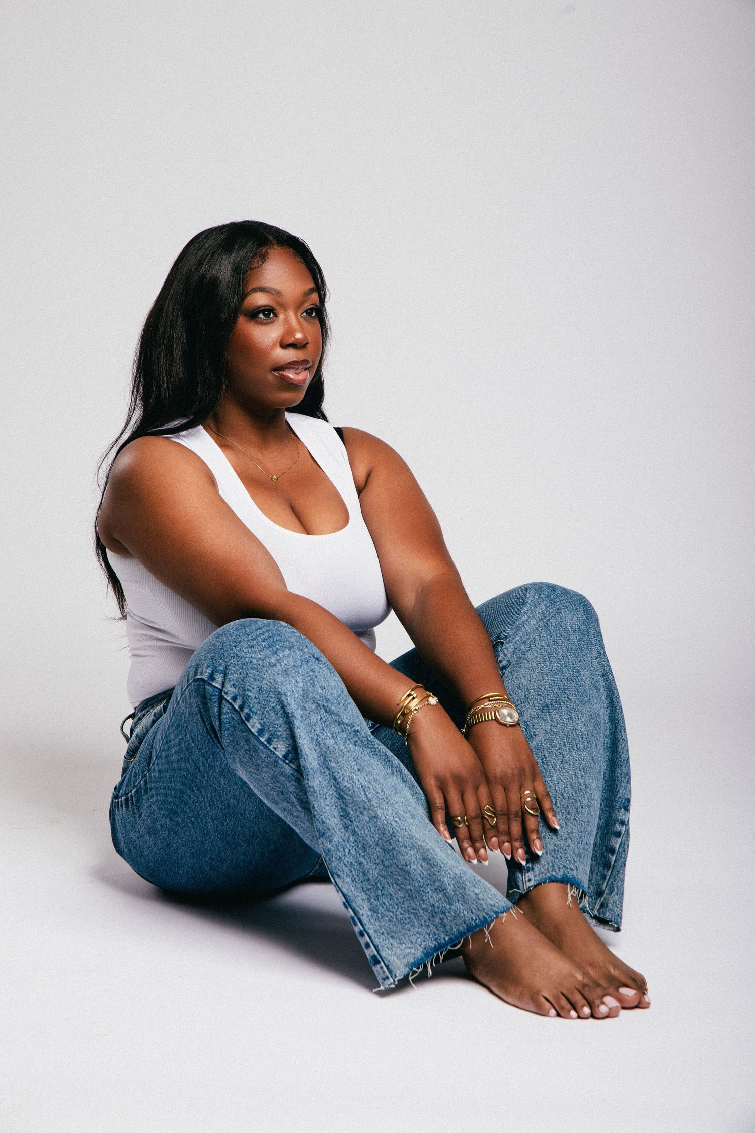 A young Black woman sits on a white floor wearing a white tank top, blue wide-leg jeans, and gold jewelry.