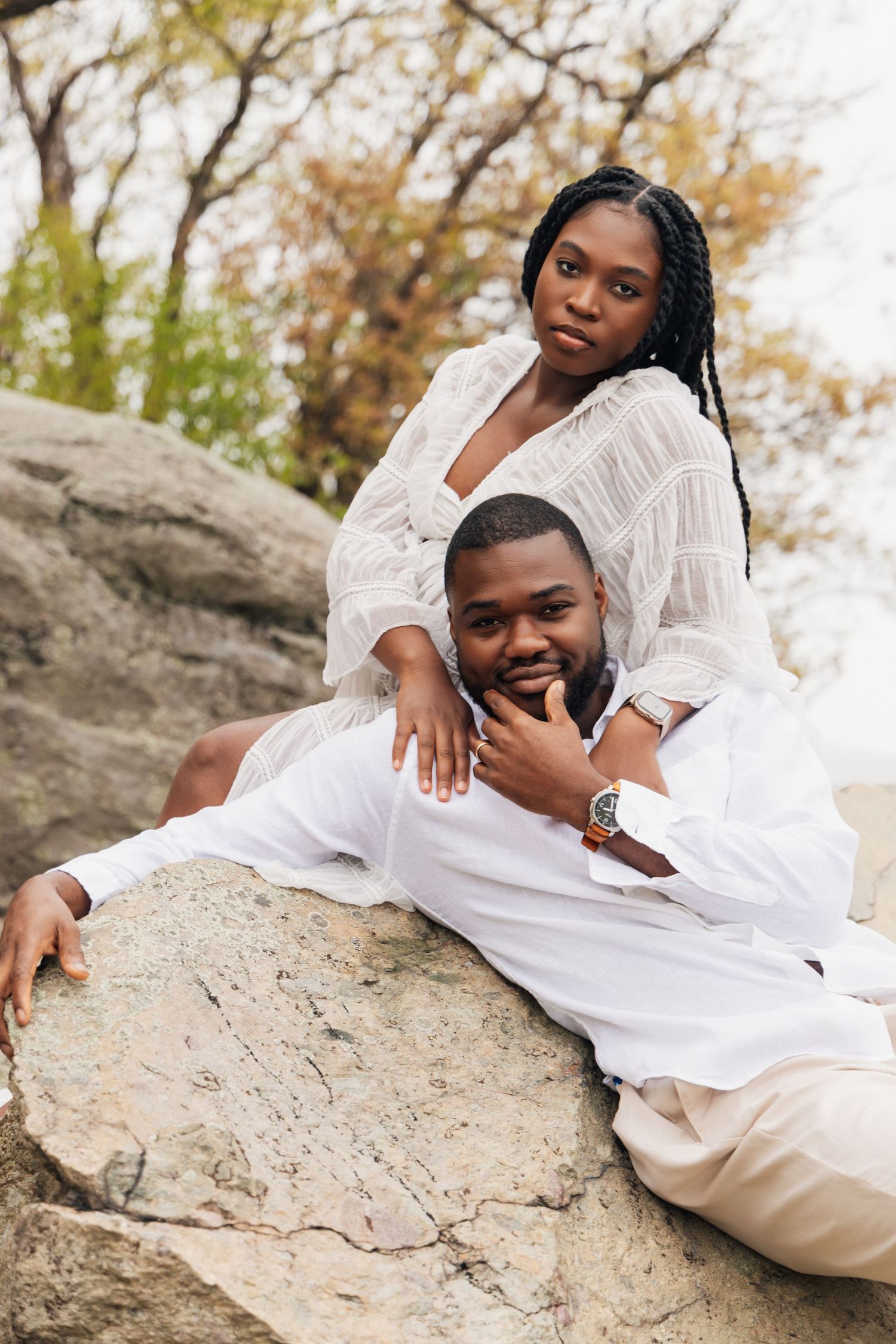 Black couple in matching white outfits posing on rocks outdoors, woman sitting above man who rests against boulder.