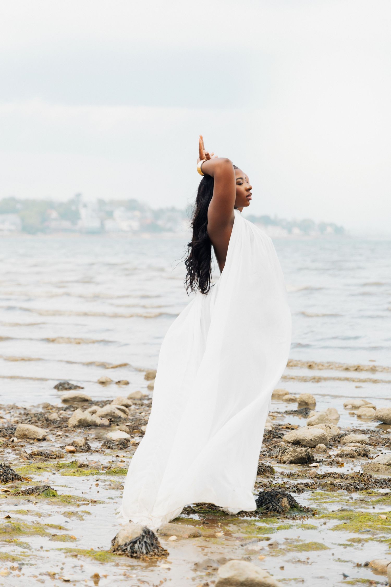 A Black woman in a flowing white fabric poses gracefully on rocky shoreline with ocean backdrop.