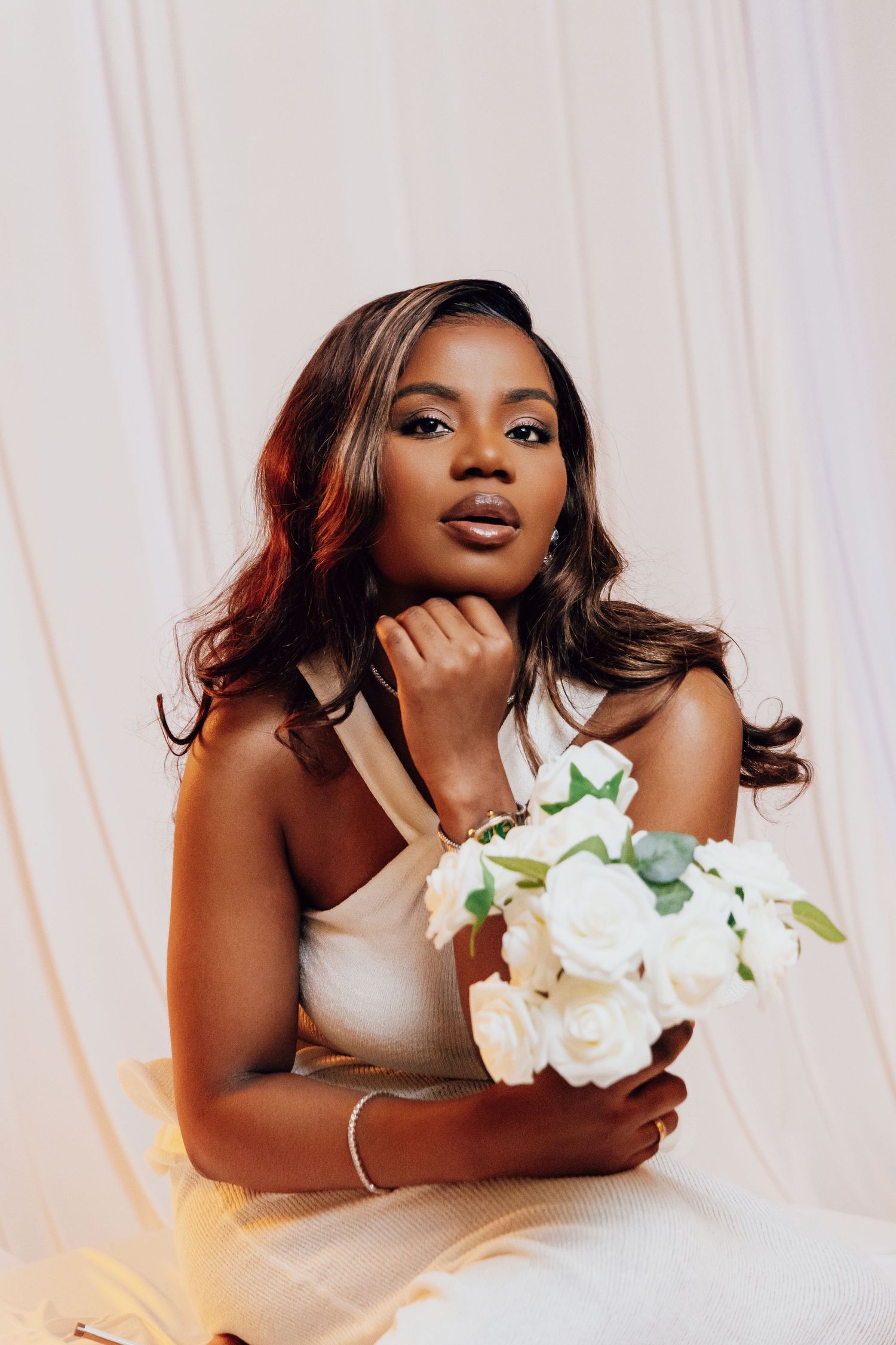 Elegant Black woman in white dress holding white roses bouquet, posing gracefully against soft draped fabric backdrop.