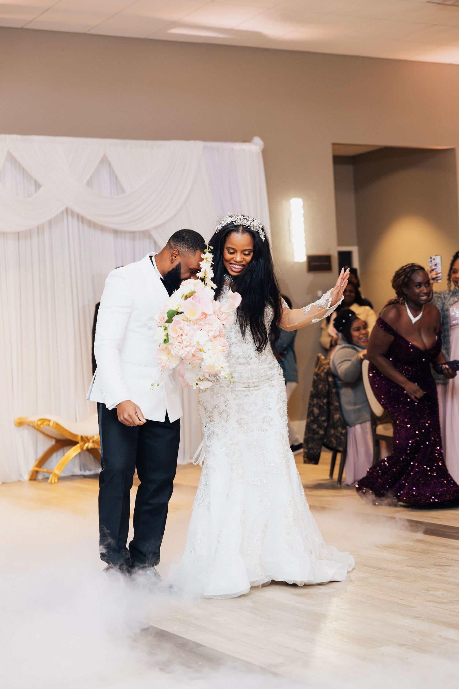 Smiling bride in beaded gown and crown walks with groom in white tuxedo through fog at wedding reception.