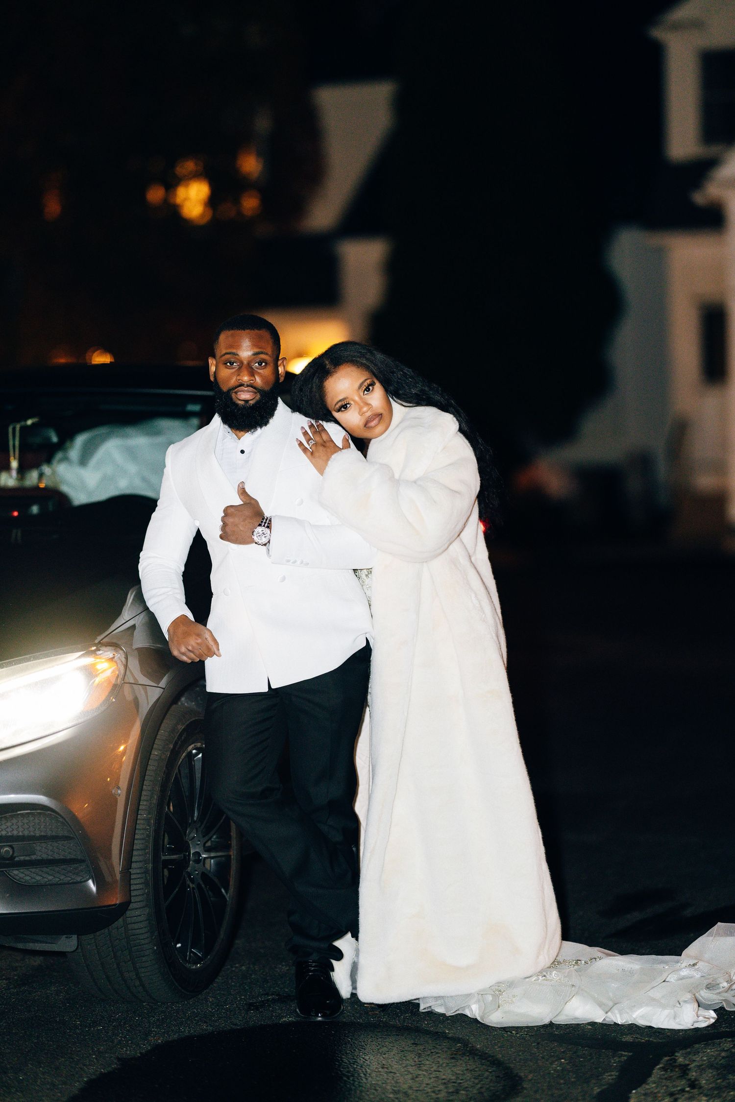 Bride in white fur coat and groom in white shirt pose together beside a car at night after wedding.