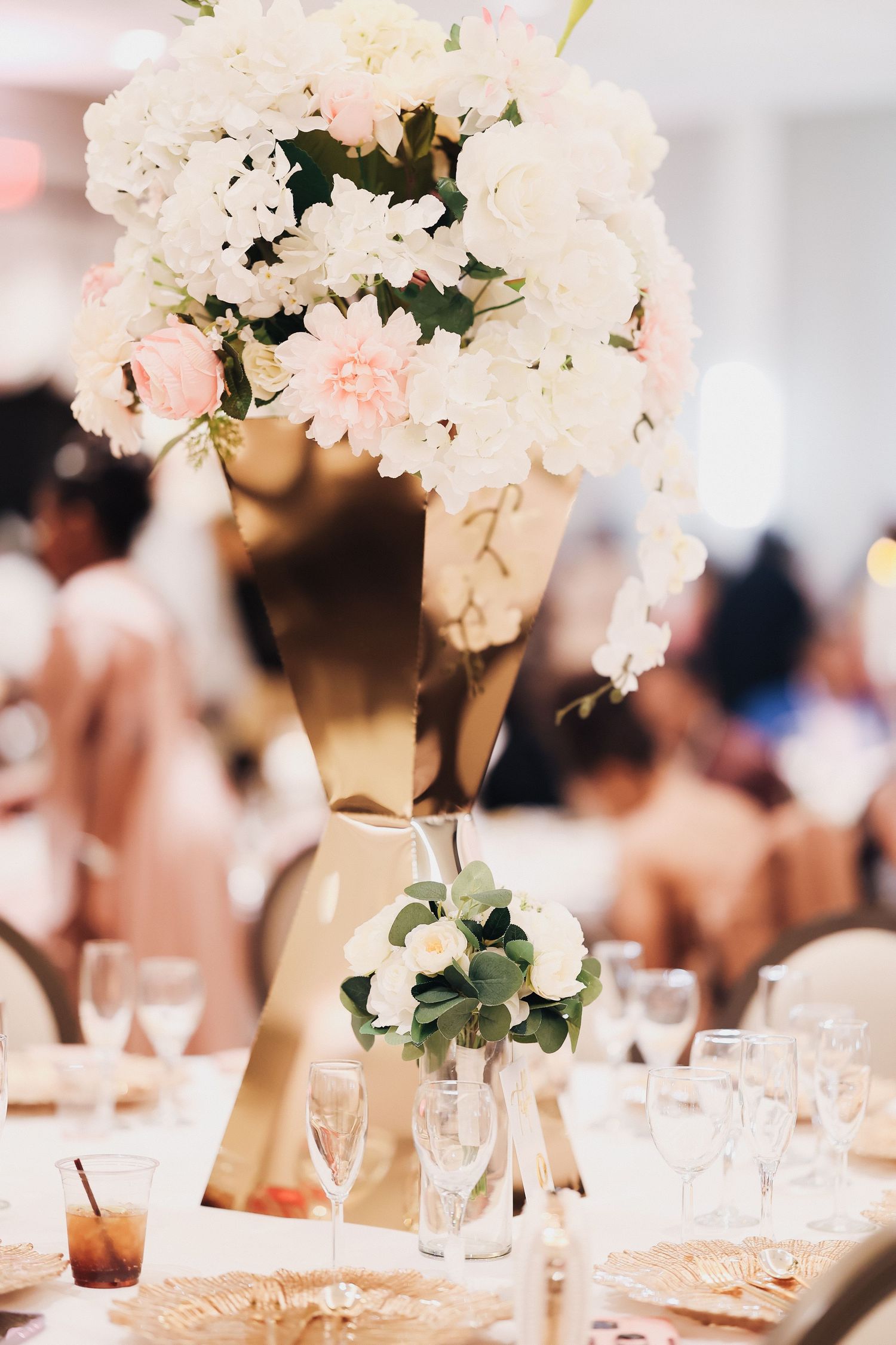 Elegant wedding reception table with tall gold vase centerpiece featuring white and pink flowers, crystal glasses, and blurred guests.